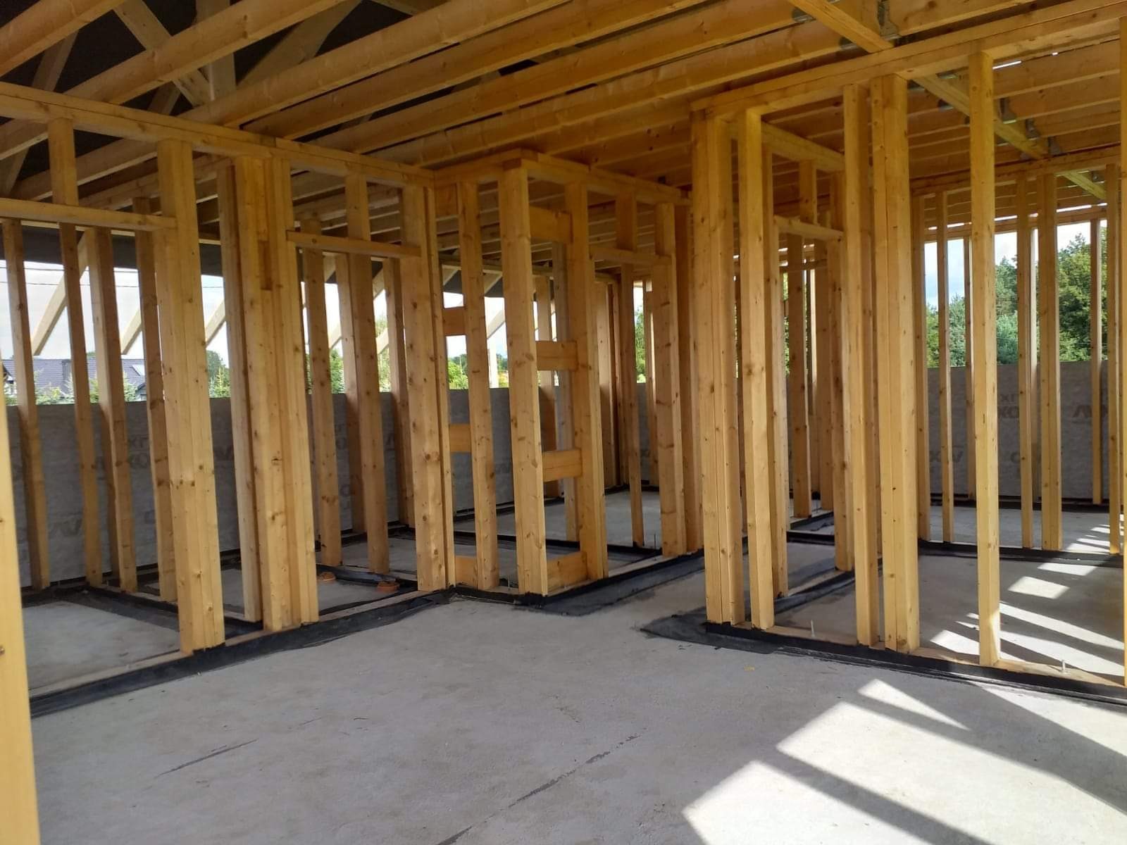 Wnętrze domu w trakcie budowy z drewnianą konstrukcją szkieletową i betonową wylewką podłogi, widoczne czarne zabezpieczenie przeciwwilgociowe u podstawy ścian.