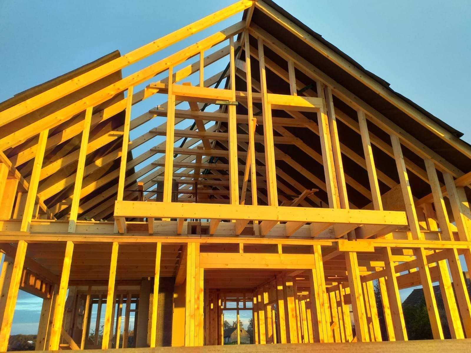 Wznoszenie konstrukcji domu z drewnianych belek w promieniach zachodzącego słońca, widoczne elementy dachu i ścian.