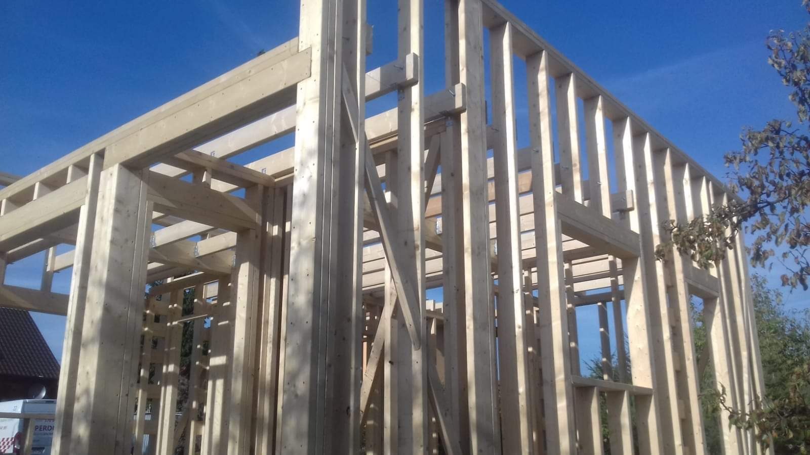 Wznoszenie drewnianej konstrukcji szkieletowej domu z widocznymi elementami łączącymi, jasne drewno na tle błękitnego nieba.