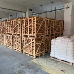 Stosy drewnianych skrzyń wypełnionych pociętym, jasnym drewnem opałowym na paletach w magazynie, obok paleta z workami foliowymi.