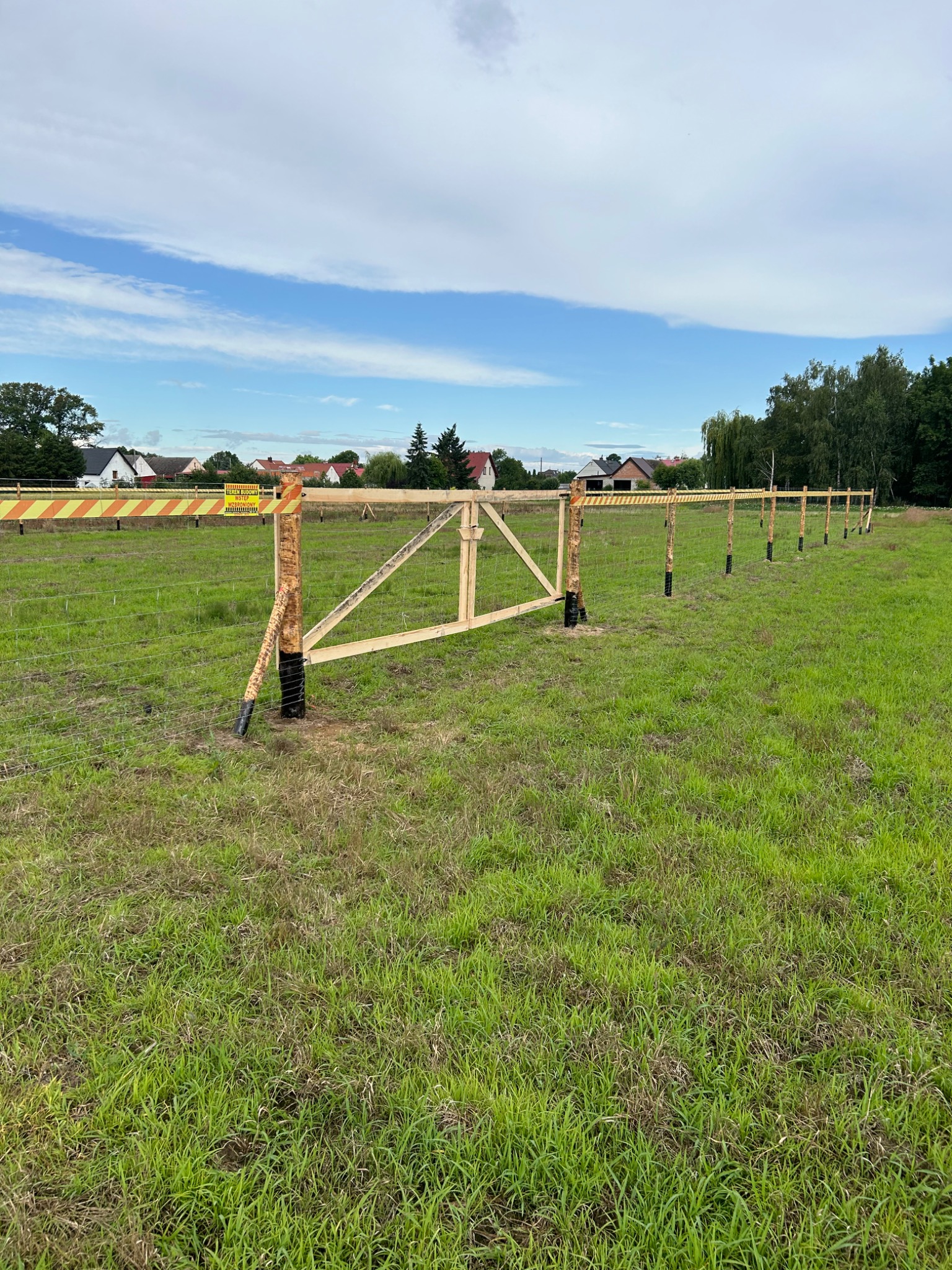 W trakcie budowy ogrodzenia z drewnianych słupków i siatki, z żółto-czarną taśmą ostrzegawczą, na tle zielonej łąki i domów.