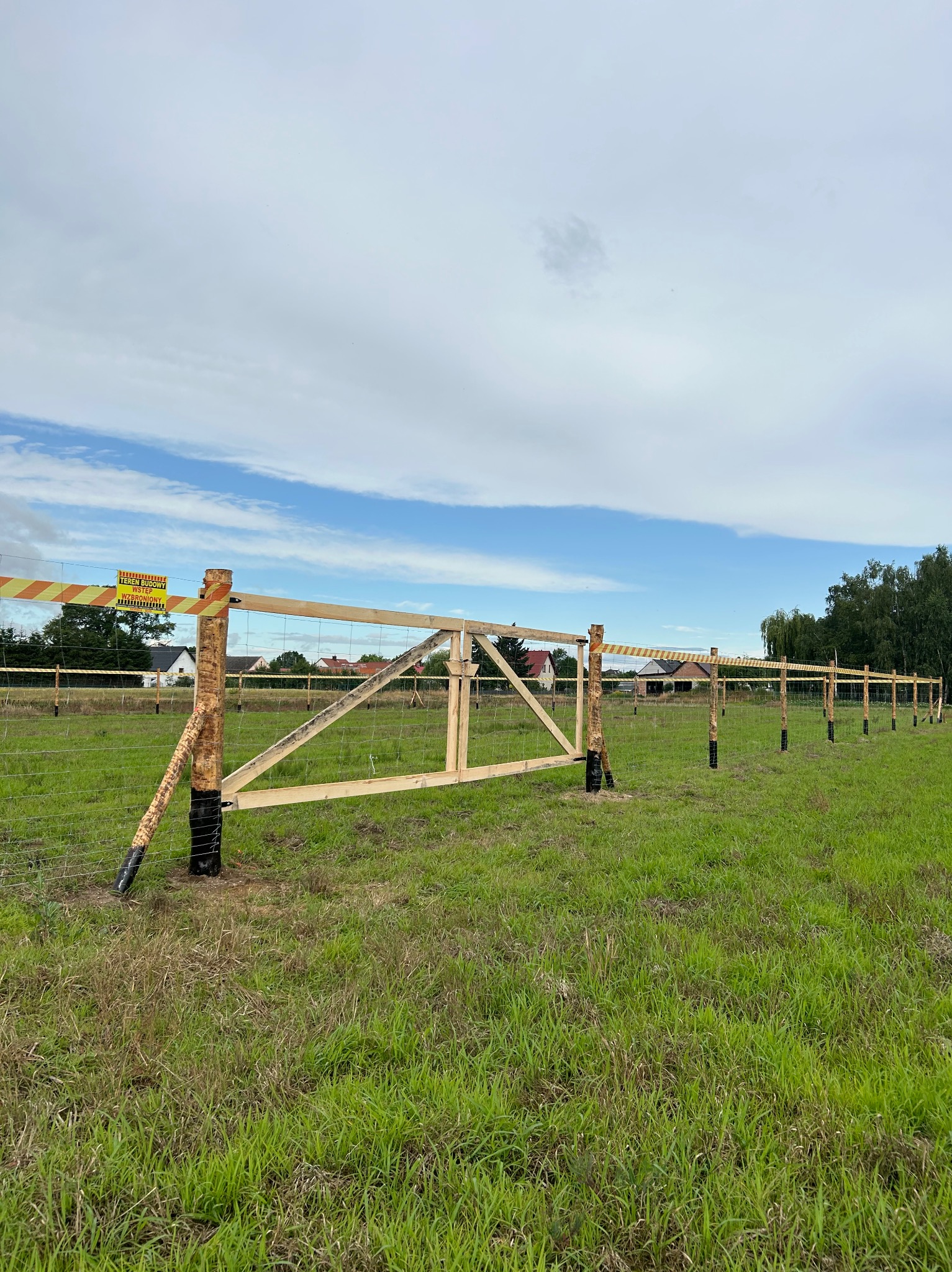 Wznoszone ogrodzenie z siatki na drewnianych słupkach, zabezpieczone taśmą ostrzegawczą, widok na rozległy teren zielony i zabudowania w tle.