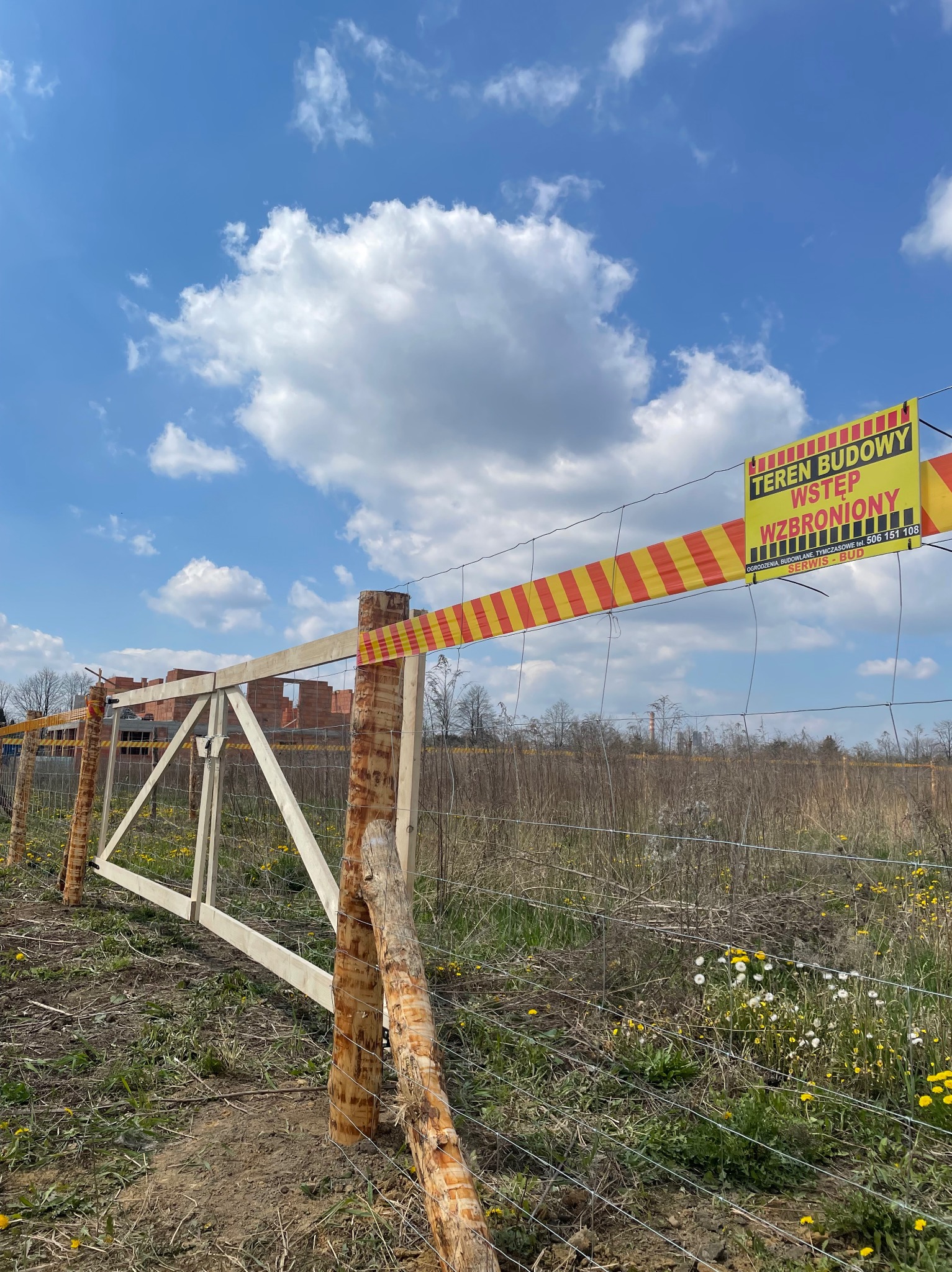 Tymczasowe ogrodzenie z siatki z drewnianymi słupkami i żółto-czerwoną taśmą ostrzegawczą na terenie budowy, w tle wznoszony budynek z czerwonej cegły i błękitne niebo z chmurami.