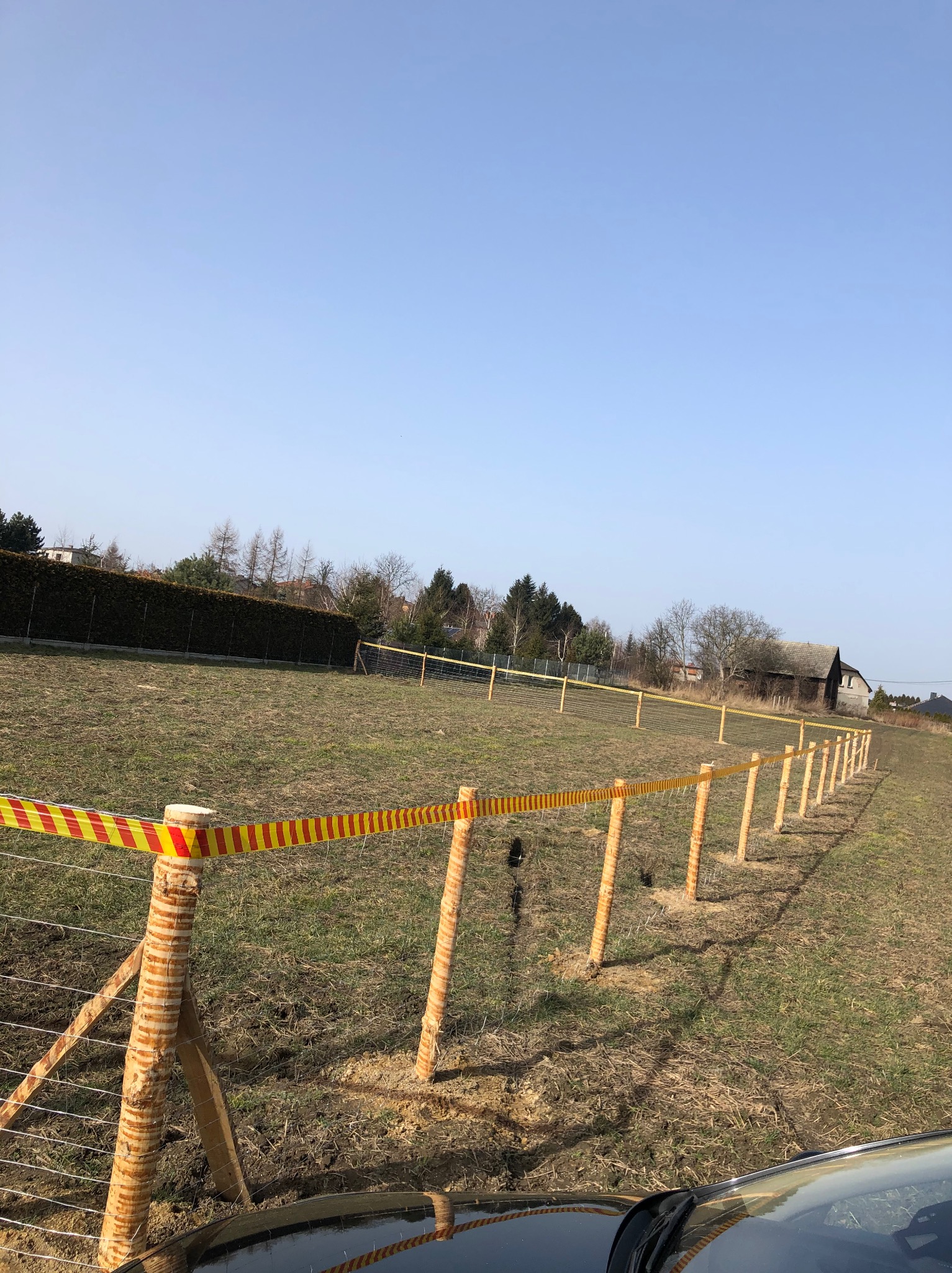 Nowo postawione ogrodzenie z siatki z drewnianymi słupkami, zabezpieczone taśmą ostrzegawczą, na tle pola i zabudowań gospodarczych.