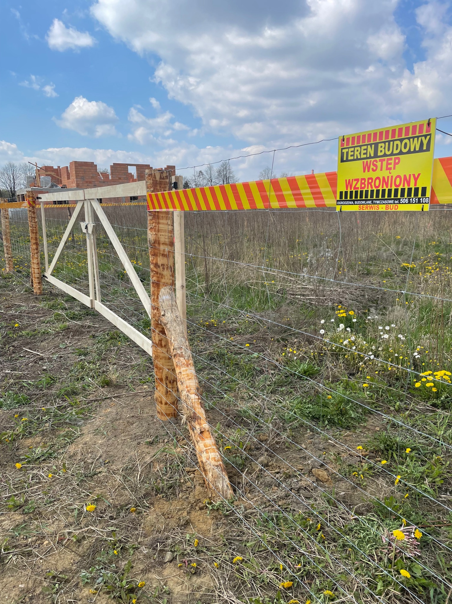 Tymczasowe ogrodzenie z siatki z drewnianą bramą na terenie budowy, żółto-czerwona taśma ostrzegawcza i tablica informacyjna, w tle niedokończony budynek z czerwonej cegły.