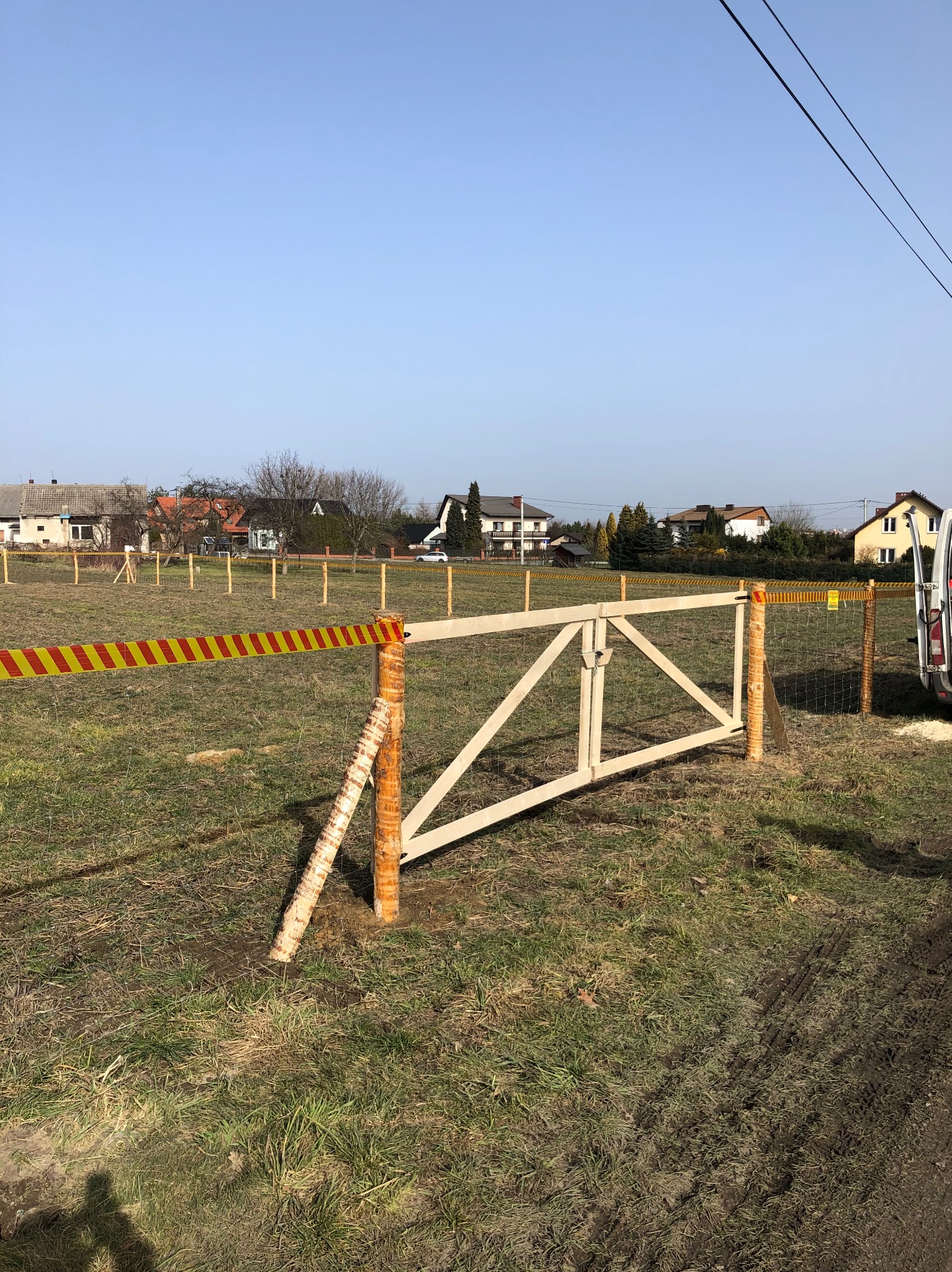 W trakcie montażu drewnianej bramy w ogrodzeniu z siatki leśnej, słupki z okorowanej brzozy, teren nierówny, widoczne domy w tle.