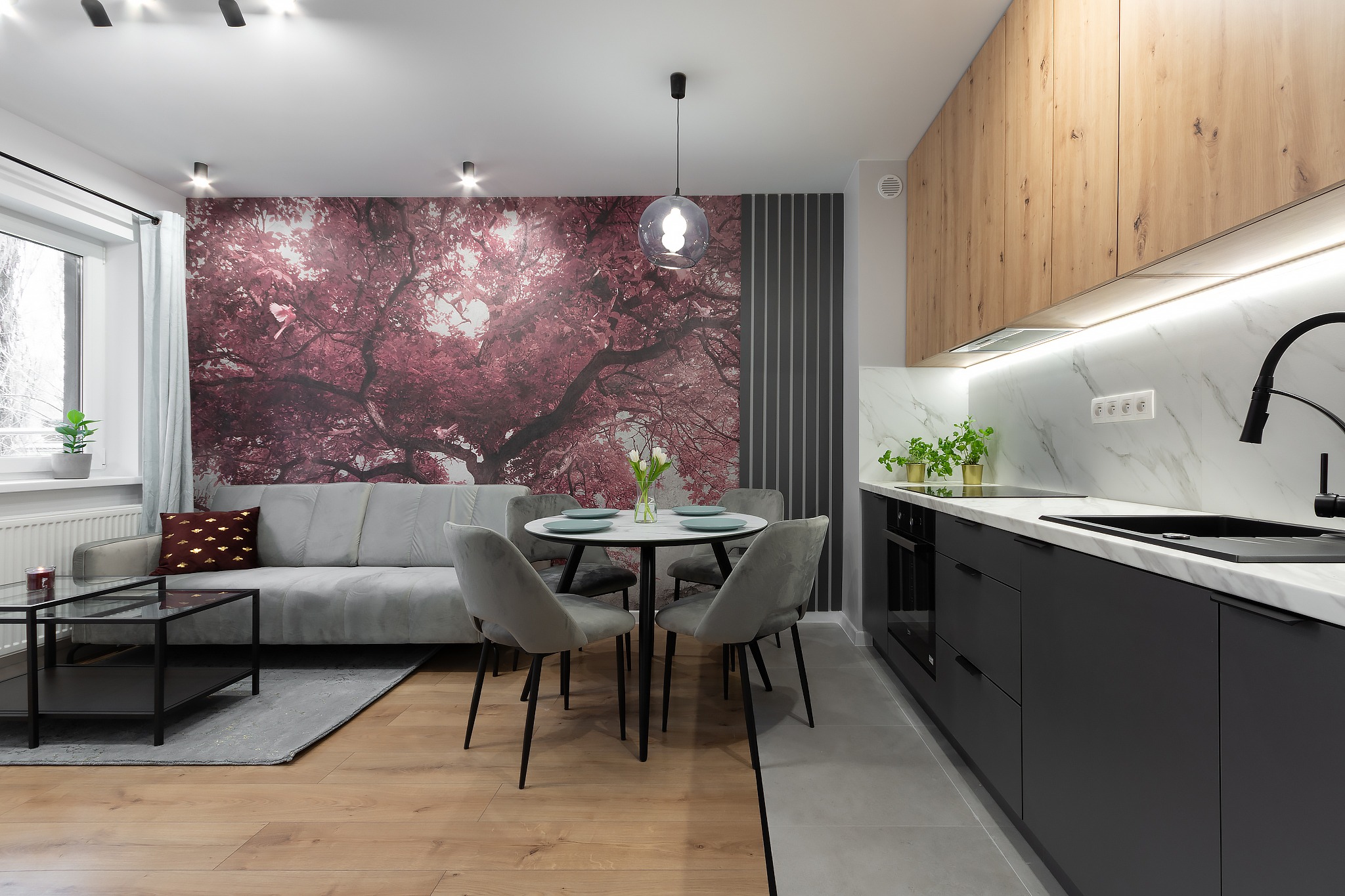 Nowoczesne wnętrze: szara sofa na tle fototapety z kwitnącym drzewem, okrągły stół z szarymi krzesłami i czarna zabudowa kuchenna z białym blatem.