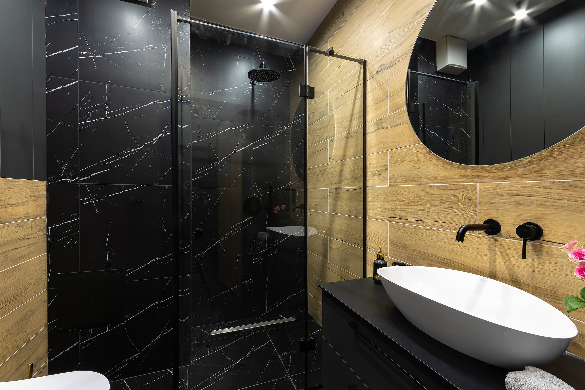 Nowoczesna łazienka z czarnymi płytkami imitującymi marmur z białymi żyłami, kabiną prysznicową typu walk-in, drewnopodobnymi płytkami na ścianie, okrągłym lustrem i białą, owalną umywalką...