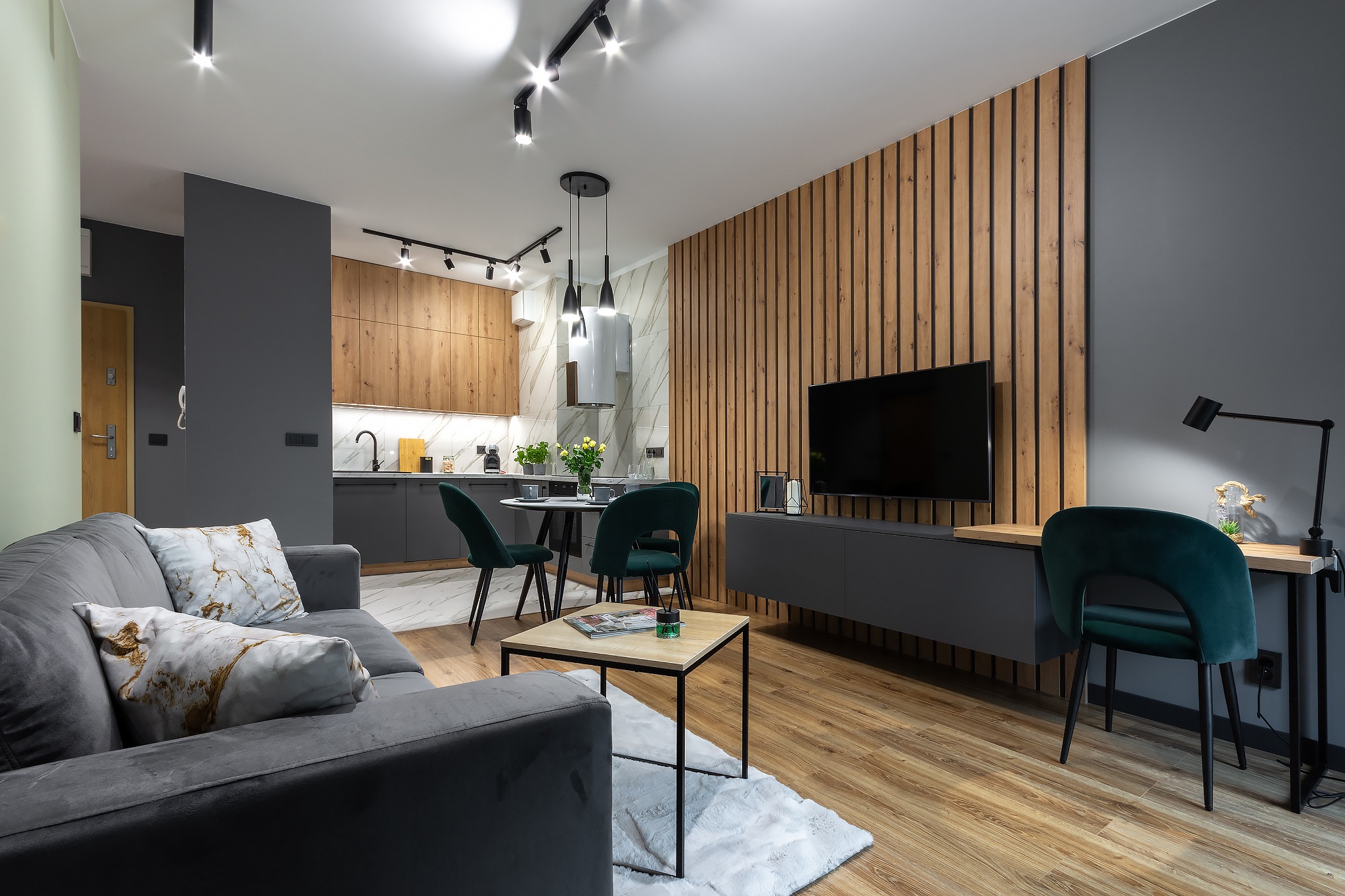 Nowoczesne wnętrze mieszkania z szarą sofą, drewnianą ścianą z lameli, aneksem kuchennym i ciemnozielonymi krzesłami, oświetlone punktowymi lampami.