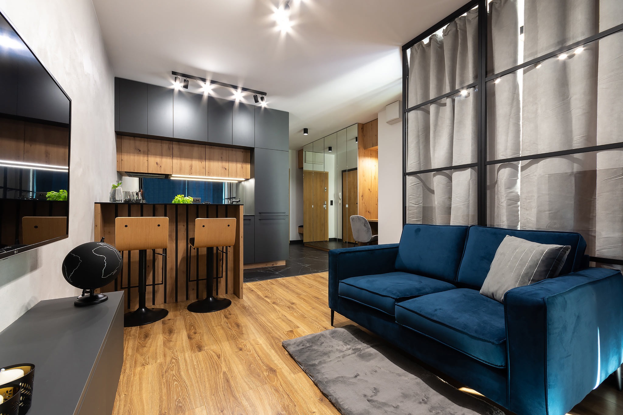 Nowoczesne wnętrze mieszkania z drewnianą podłogą, niebieską sofą i barkiem z hokerami, oświetlone reflektorami sufitowymi. Widoczny fragment kuchni z szarymi szafkami i drewnianymi elementami...