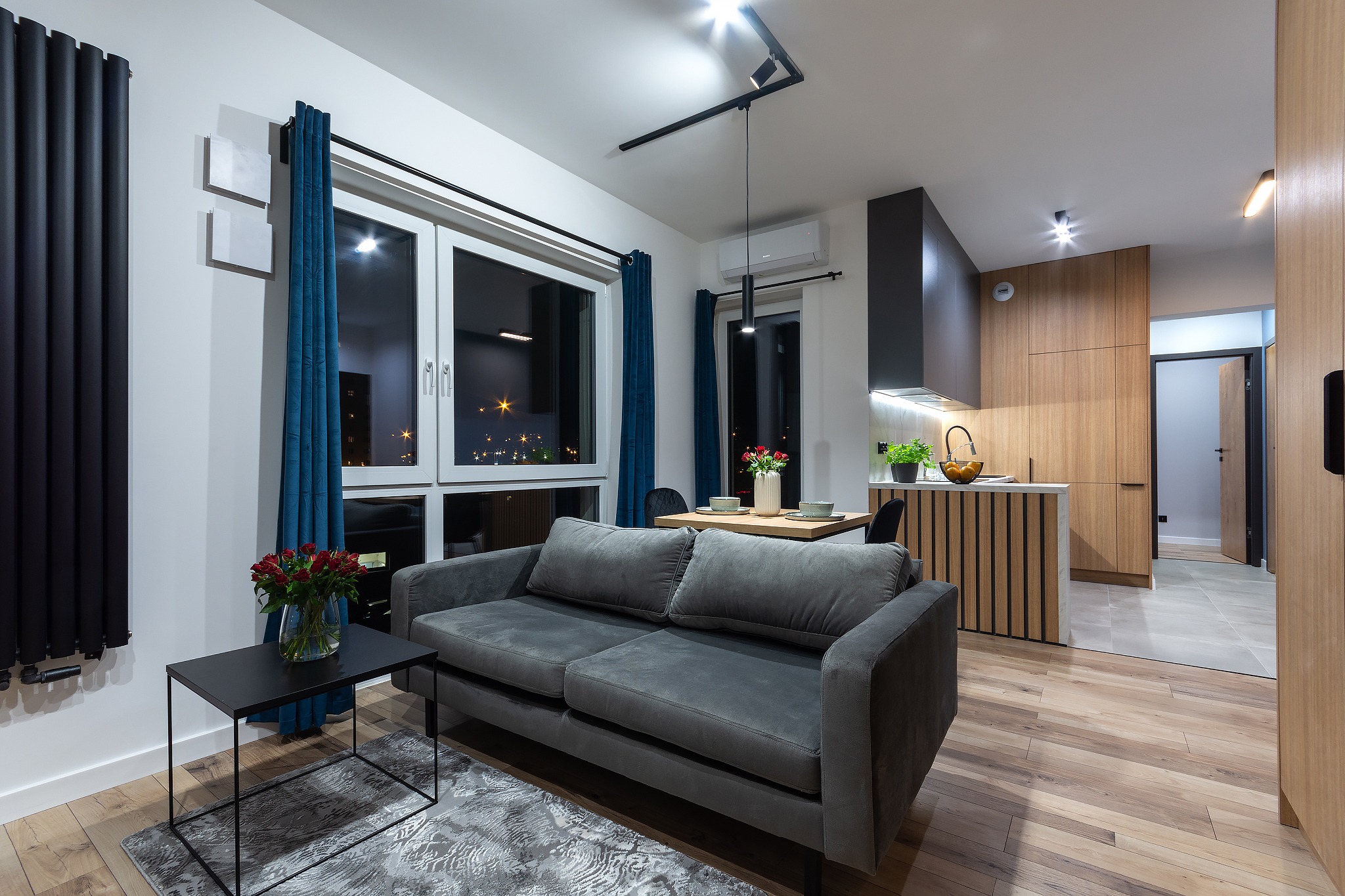 Nowoczesne wnętrze mieszkania z drewnianą podłogą, szarą sofą, czarnym grzejnikiem panelowym i otwartą kuchnią z drewnianymi szafkami, widok nocnego miasta przez okno.