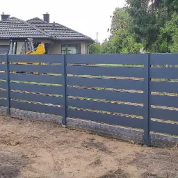 Nowoczesne szare ogrodzenie panelowe z poziomych elementów, widok z zewnątrz posesji, w tle dom i żółta koparka.