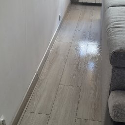 WMClean Usługi Sprzątania WERONIKA MIERZWA-KLIMCZAK - Błyszcząca, świeżo umyta podłoga z paneli imitujących drewno, widok z niskiej perspektywy, fragment sofy w tle, ściana i grzejnik.