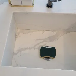 Biały, kwadratowy zlew wbudowany w blat z marmurowym wzorem, z zieloną gąbką leżącą na odpływie.