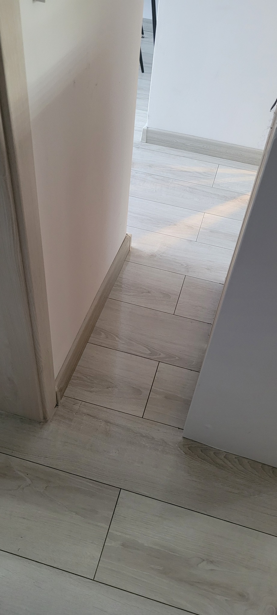 Fragment podłogi z jasnych płytek imitujących drewno, widoczna przy listwach przypodłogowych i futrynie drzwi w jasnych odcieniach.