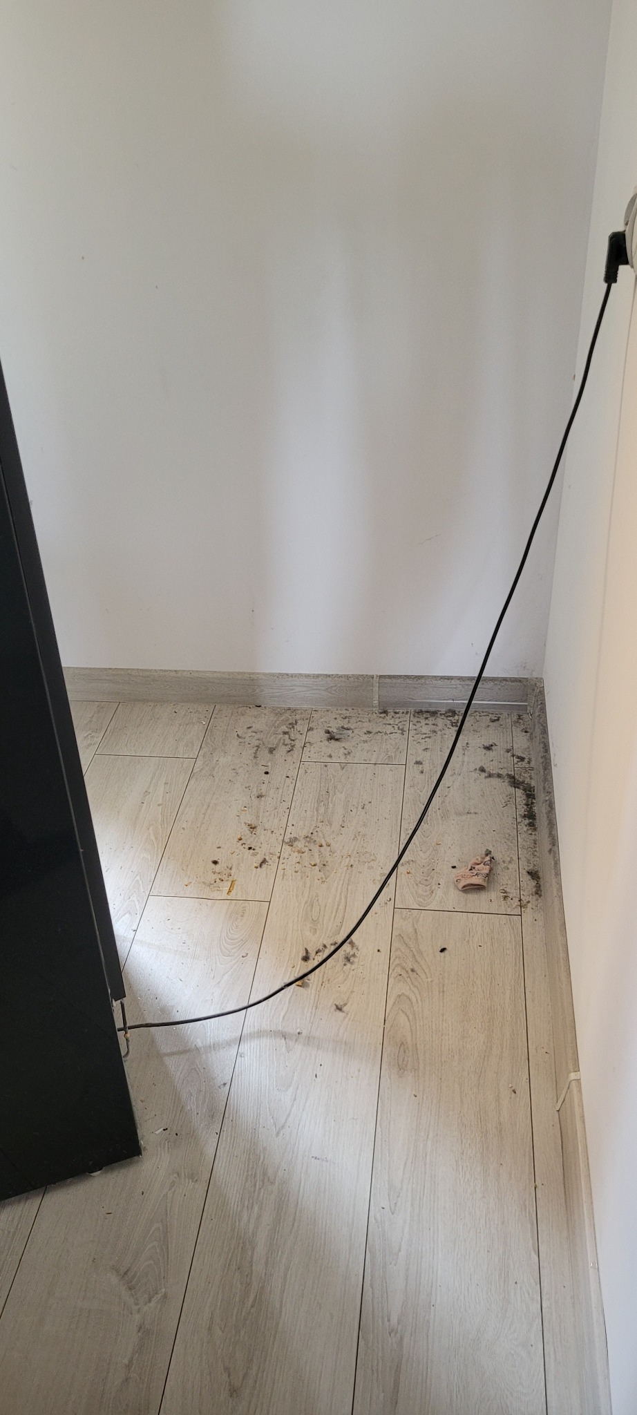Podłoga z paneli drewnopodobnych, widoczny brud, kurz i resztki po remoncie w rogu pomieszczenia, czarny kabel zasilający biegnący wzdłuż ściany.