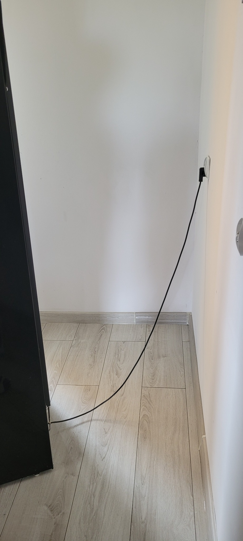 Kabel zasilający podłączony do gniazdka na białej ścianie, leżący na jasnej podłodze w rogu pokoju, obok czarnego mebla.