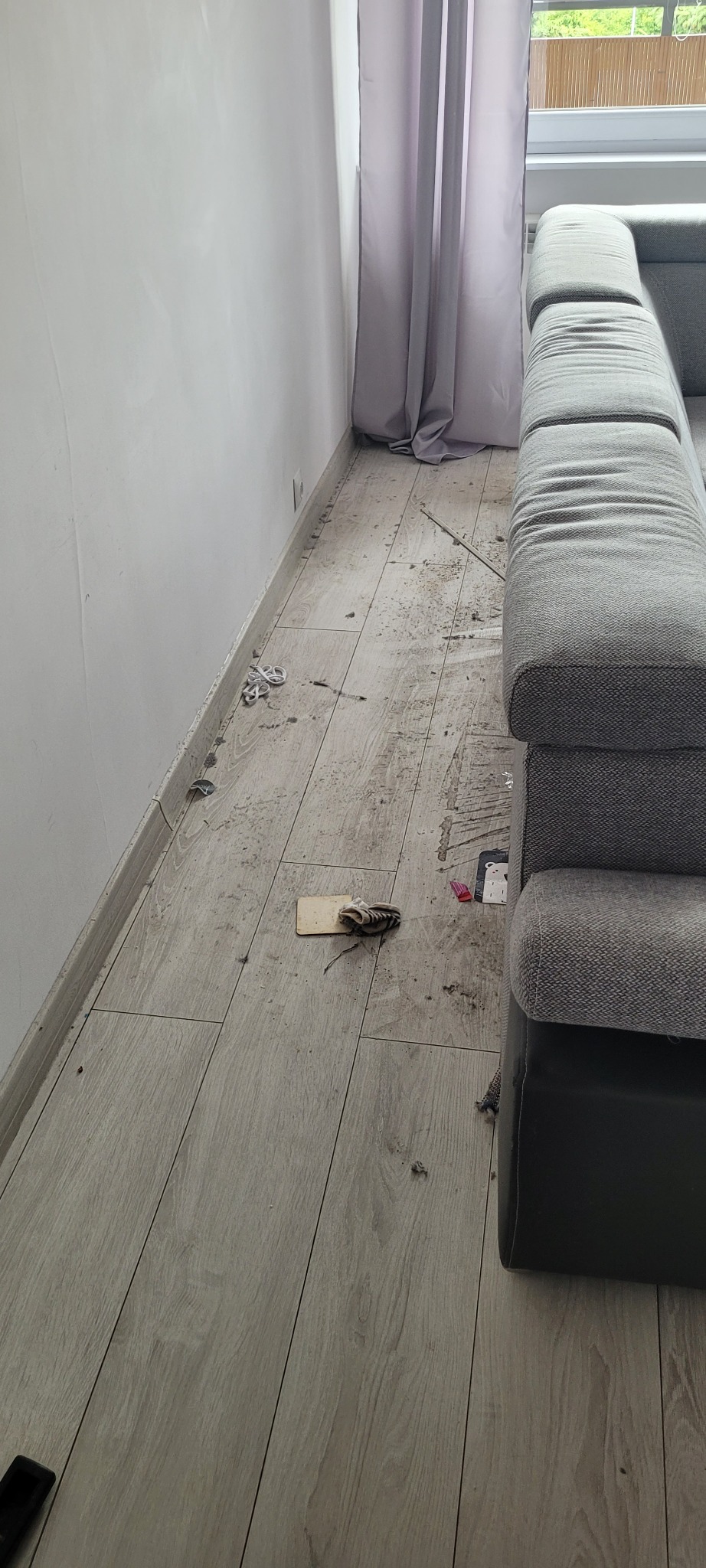 Podłoga z paneli po remoncie, widoczne drobne zabrudzenia, resztki materiałów budowlanych, obok szara sofa i zasłony w oknie.
