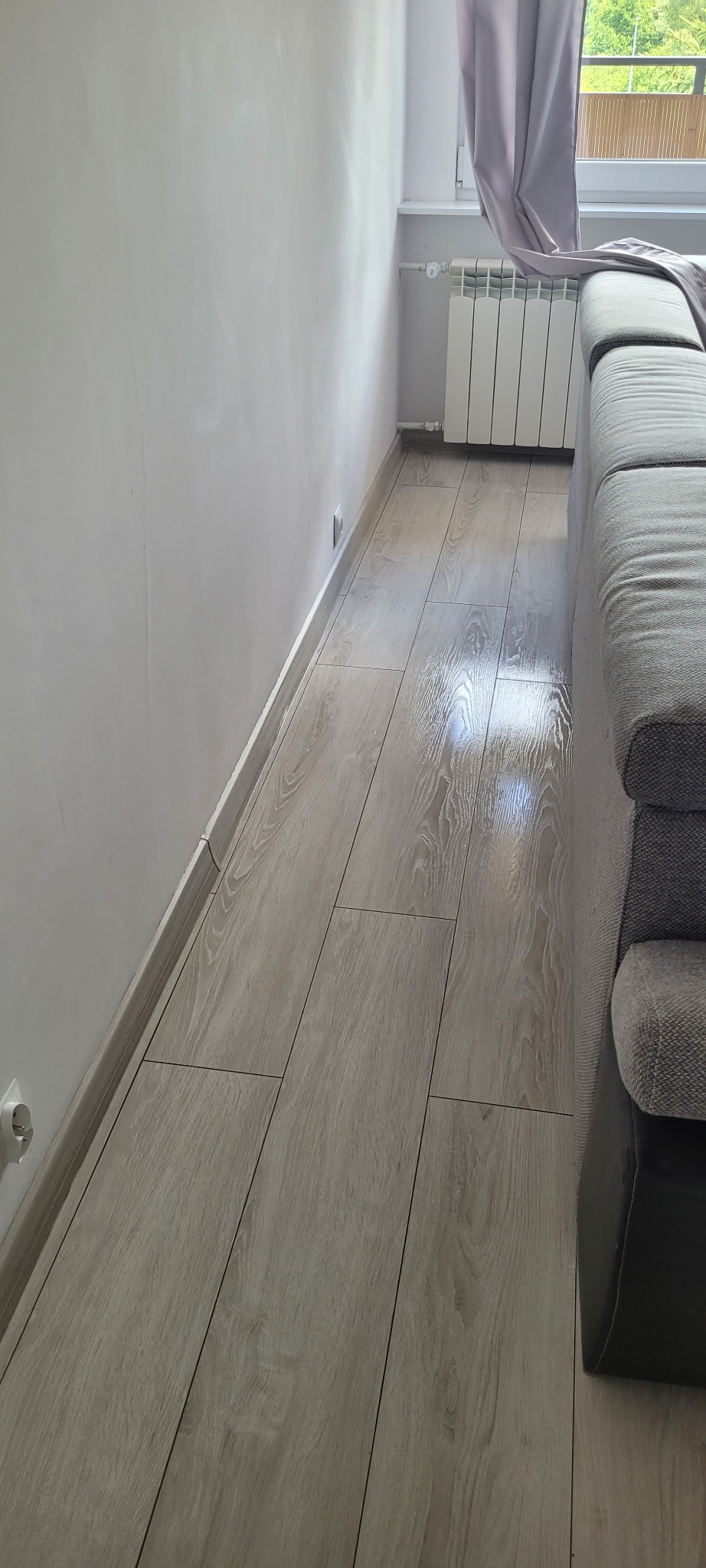 Błyszcząca, świeżo umyta podłoga z paneli imitujących drewno, widok z niskiej perspektywy, fragment sofy w tle, ściana i grzejnik.
