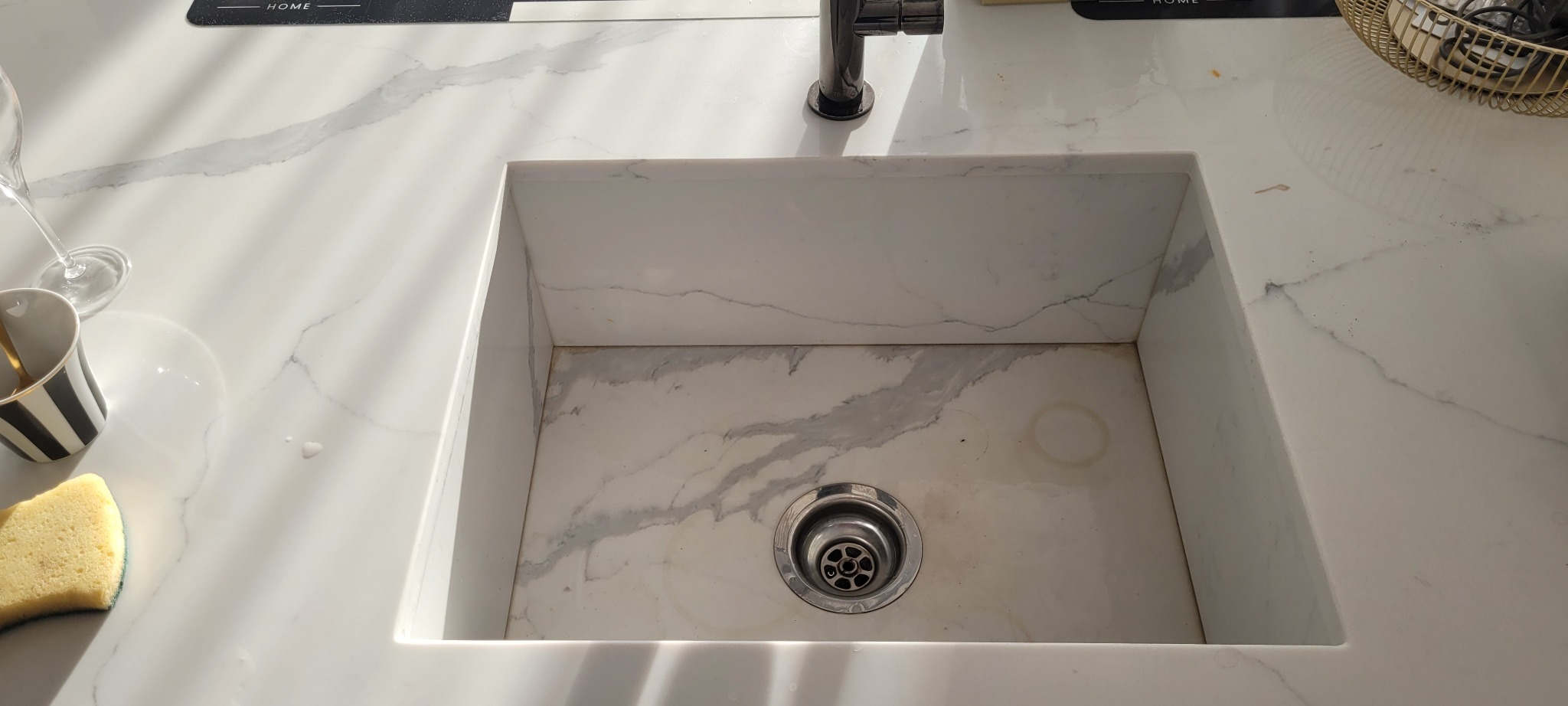 Marmurowy zlew z widocznymi śladami użytkowania, obok gąbka do mycia naczyń, kubek w paski i kieliszek do wina.