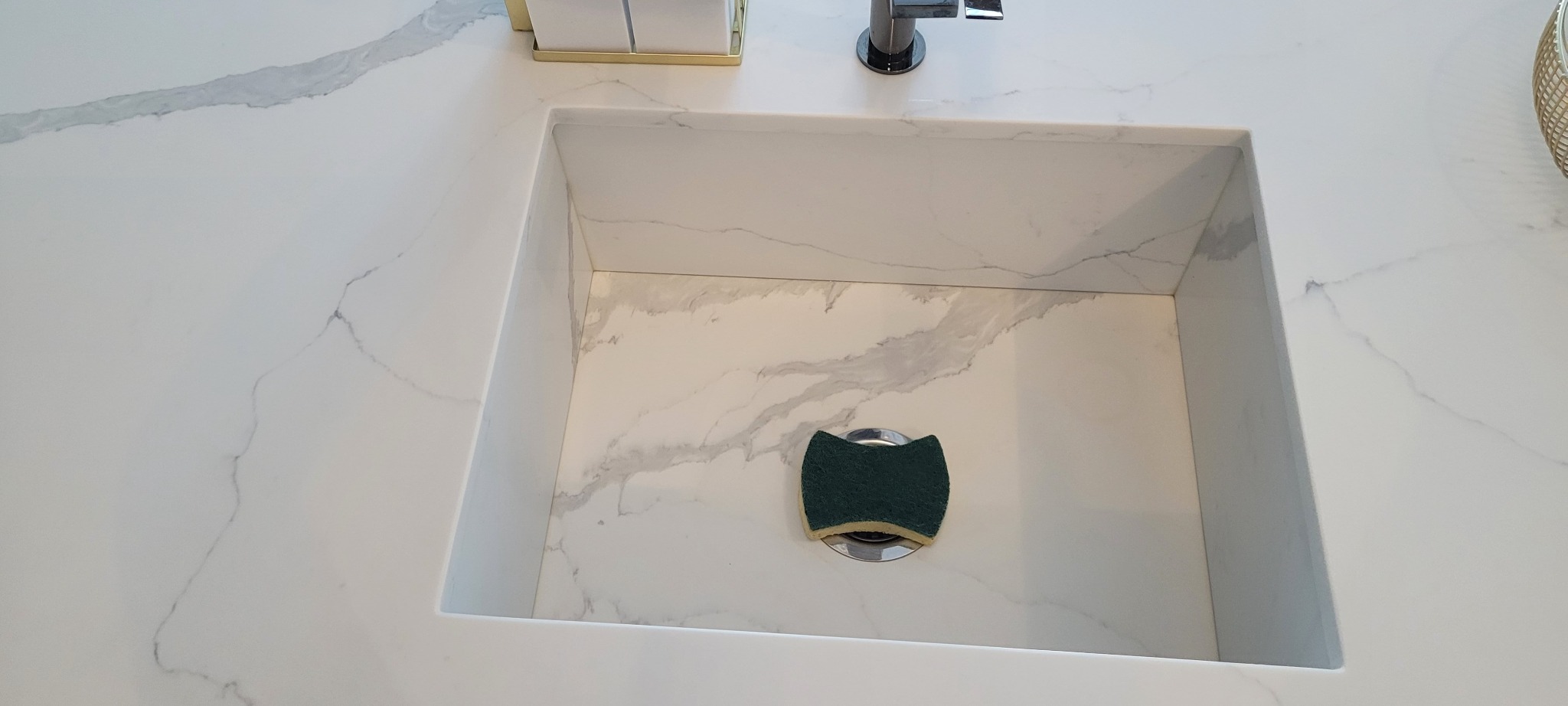 Biały, kwadratowy zlew wbudowany w blat z marmurowym wzorem, z zieloną gąbką leżącą na odpływie.