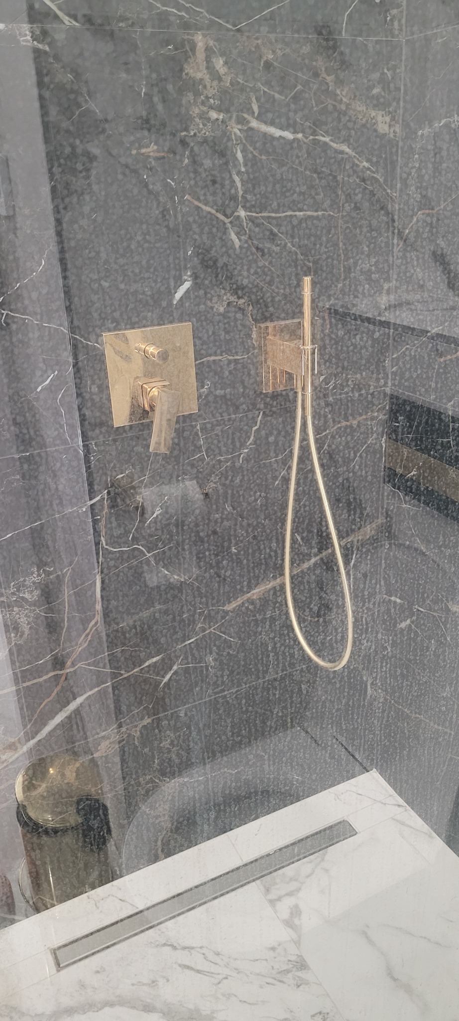 Szczegółowe ujęcie kabiny prysznicowej z czarnymi marmurowymi ścianami, złotą armaturą i odpływem liniowym w białej marmurowej podłodze, widoczne ślady po wodzie na szybie.