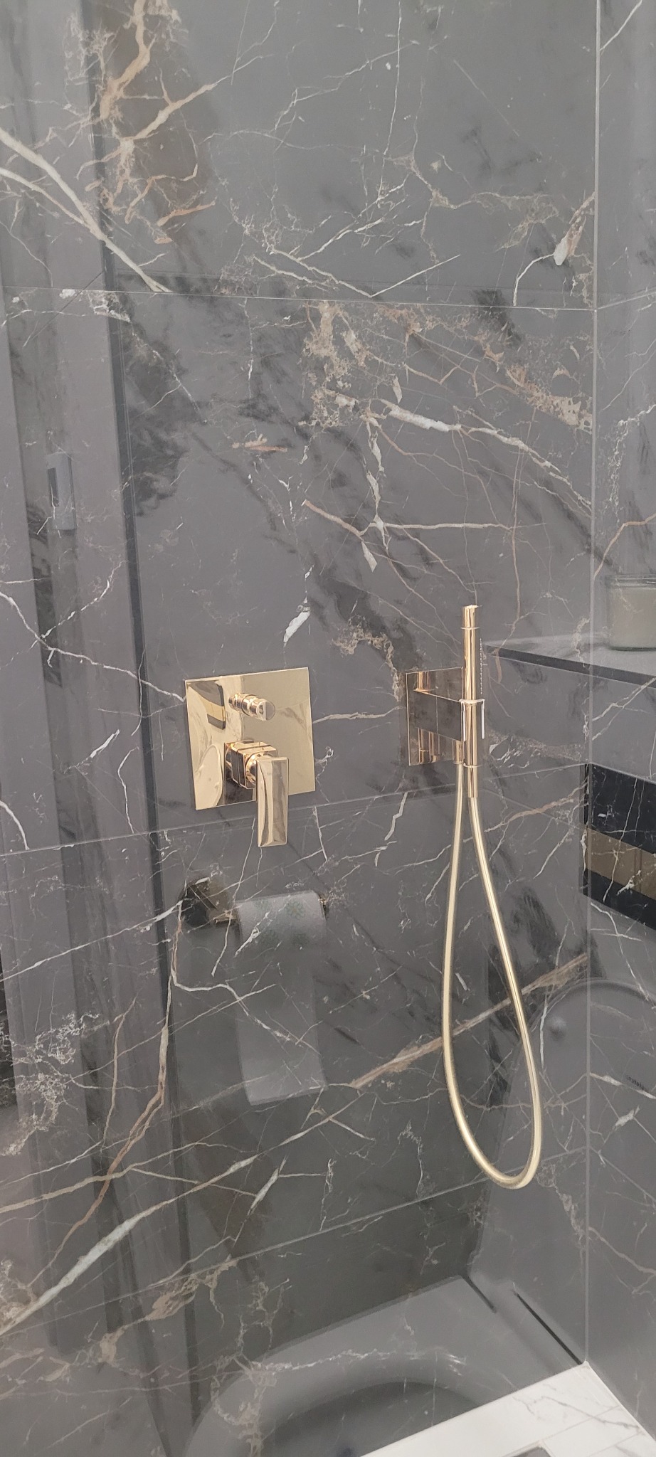 Błyszcząca, czarna marmurowa ściana kabiny prysznicowej z złotą armaturą: bateria i słuchawka prysznicowa.