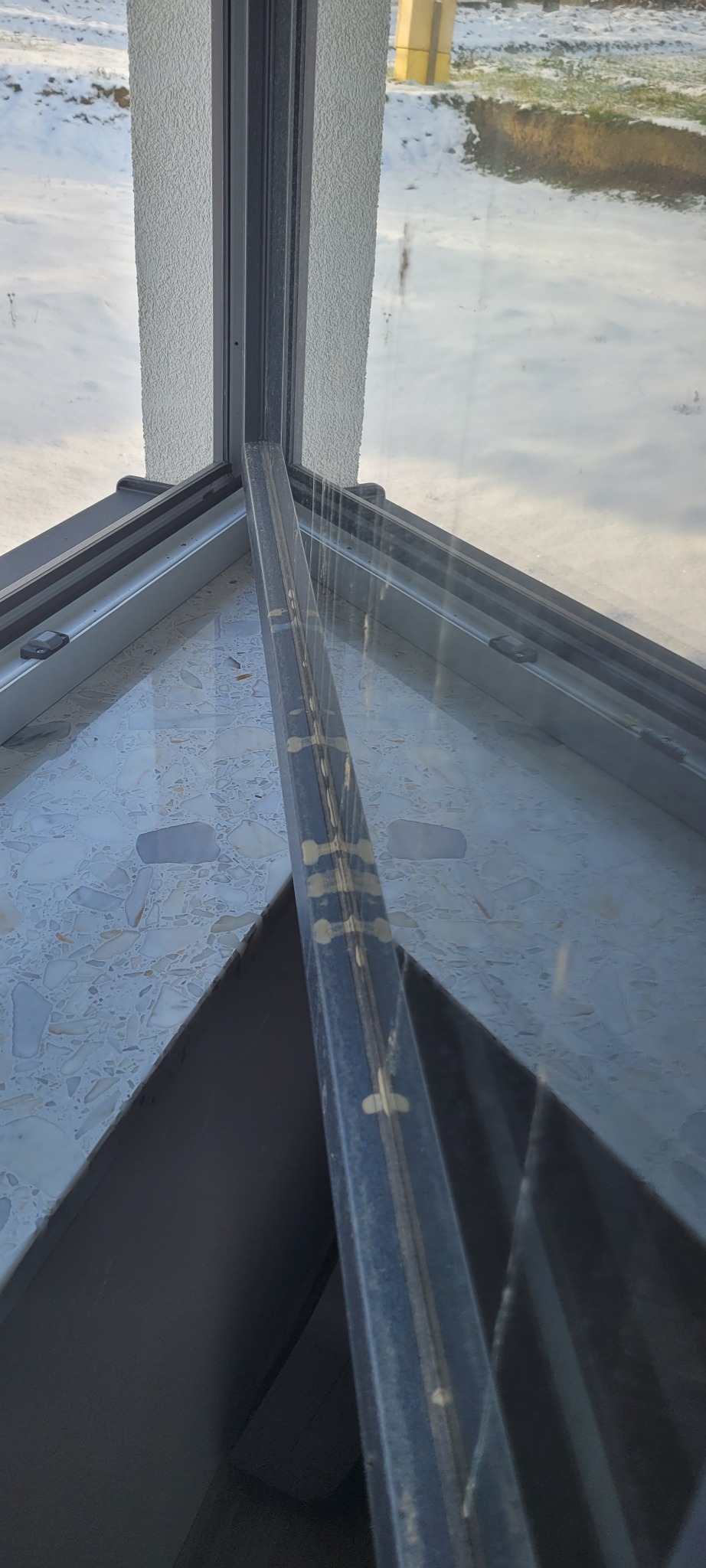 Brudna, szara rama okienna z widocznymi plamami i smugami po czyszczeniu, z zewnątrz widok na zaśnieżony teren i budynek w tle.