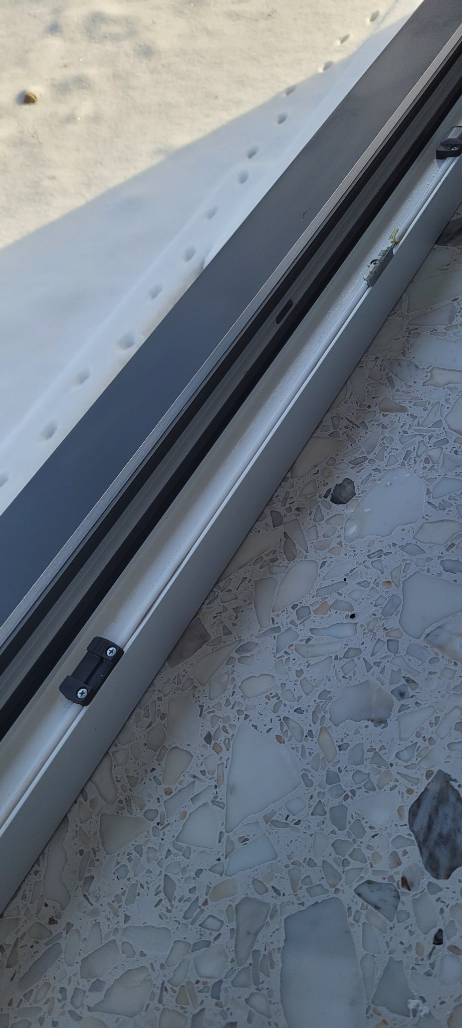 Fragment okna PCV z widocznymi śladami zwierząt na śniegu za oknem i wzorzystą posadzką wewnątrz.