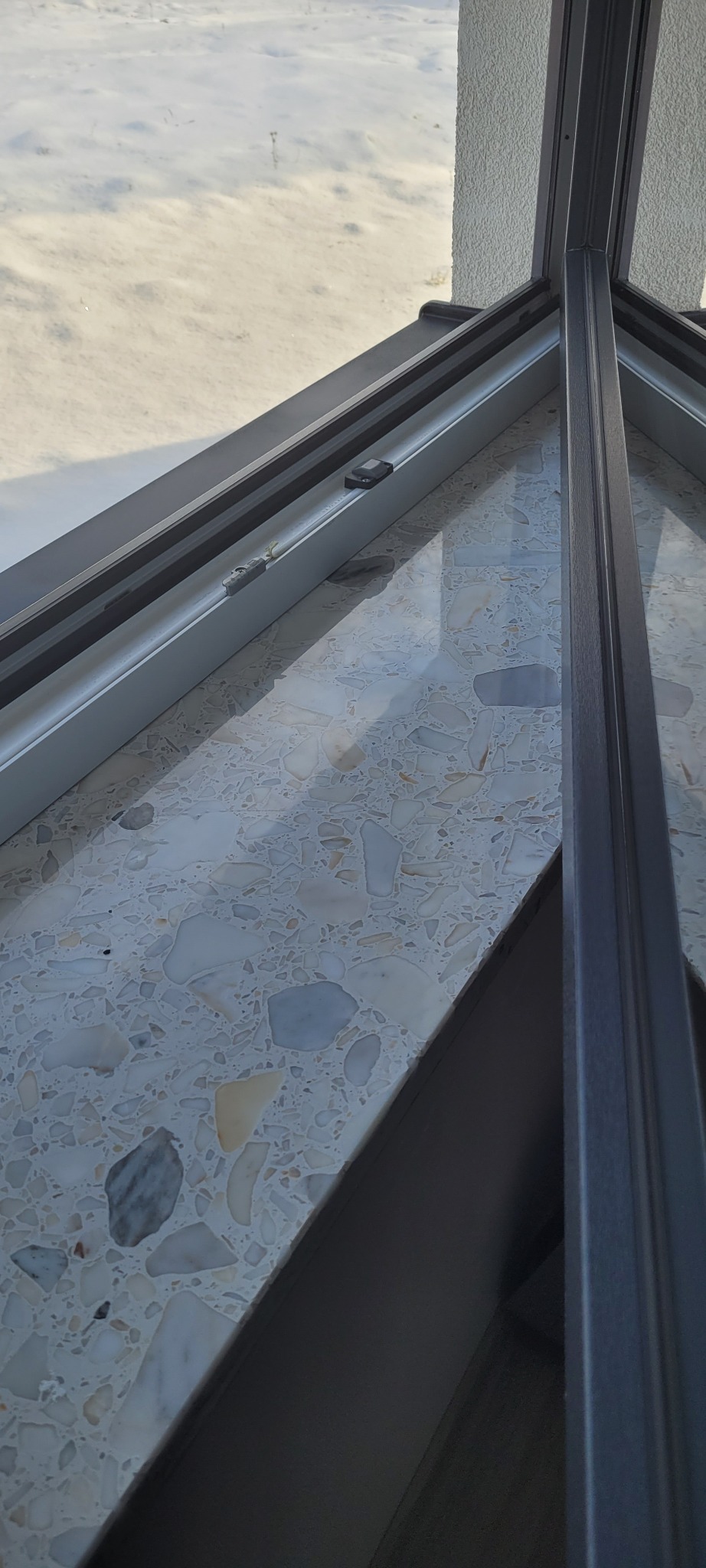 Wnętrze okna dachowego z widokiem na zaśnieżony dach, widoczne elementy okna i parapet wykończony terazzo.