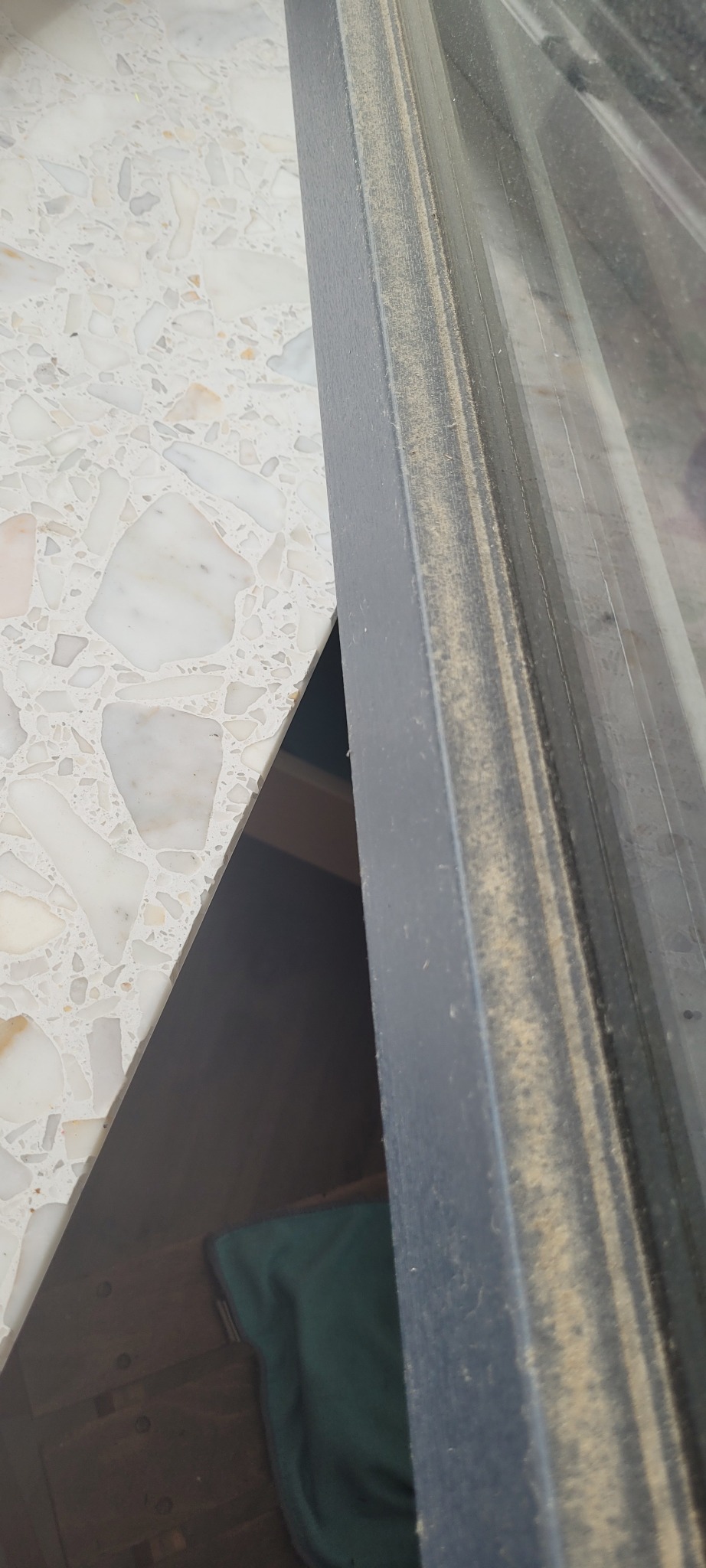 Zakurzona rama okienna z widocznymi smugami brudu i blatem stołu z wzorem lastryko w tle.