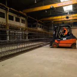 Operator w pomarańczowej kamizelce transportuje wózkiem widłowym długi element z siatki z prętów na hali produkcyjnej z suwnicą Konecranes