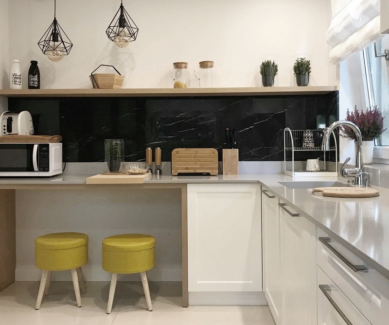 Jasna kuchnia z czarnym marmurowym backsplash, drewnianą półką z dekoracjami i dwoma żółtymi stołkami barowymi na drewnianych nogach.