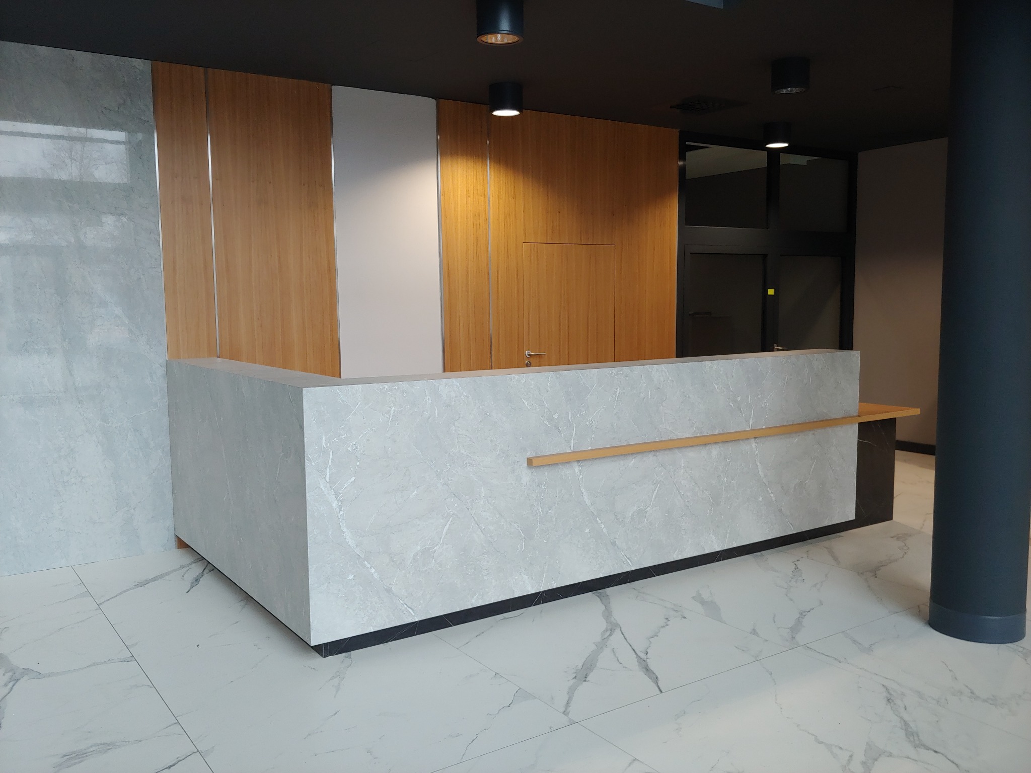 Recepcja z blatem imitującym marmur i drewnianą zabudową w Bielinach, oświetlona punktowymi lampami sufitowymi, widoczna na tle szklanych drzwi.