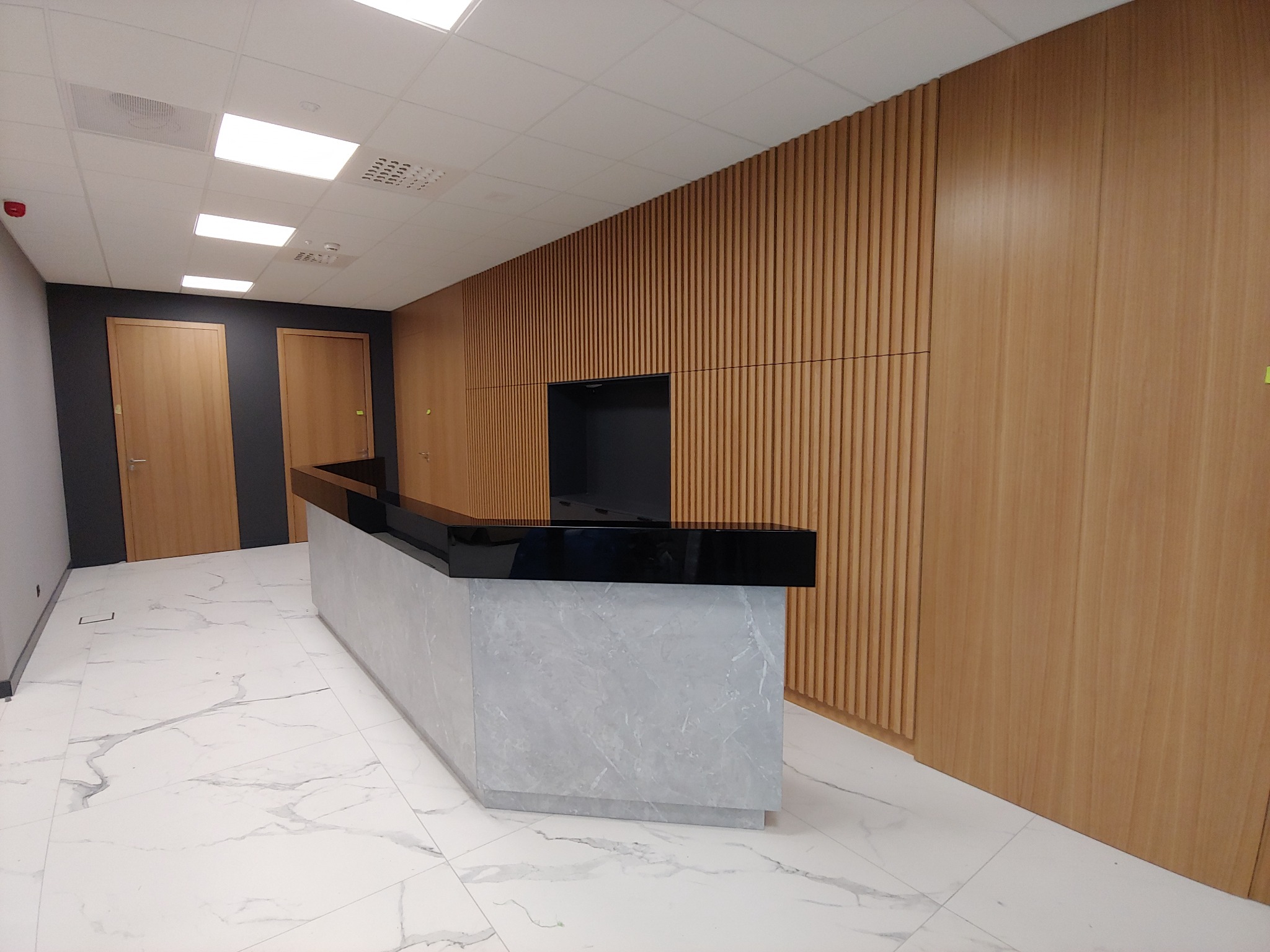 Recepcja biurowa z marmurowym blatem i drewnianą ścianą z pionowymi panelami, oświetlona światłem z sufitu kasetonowego.
