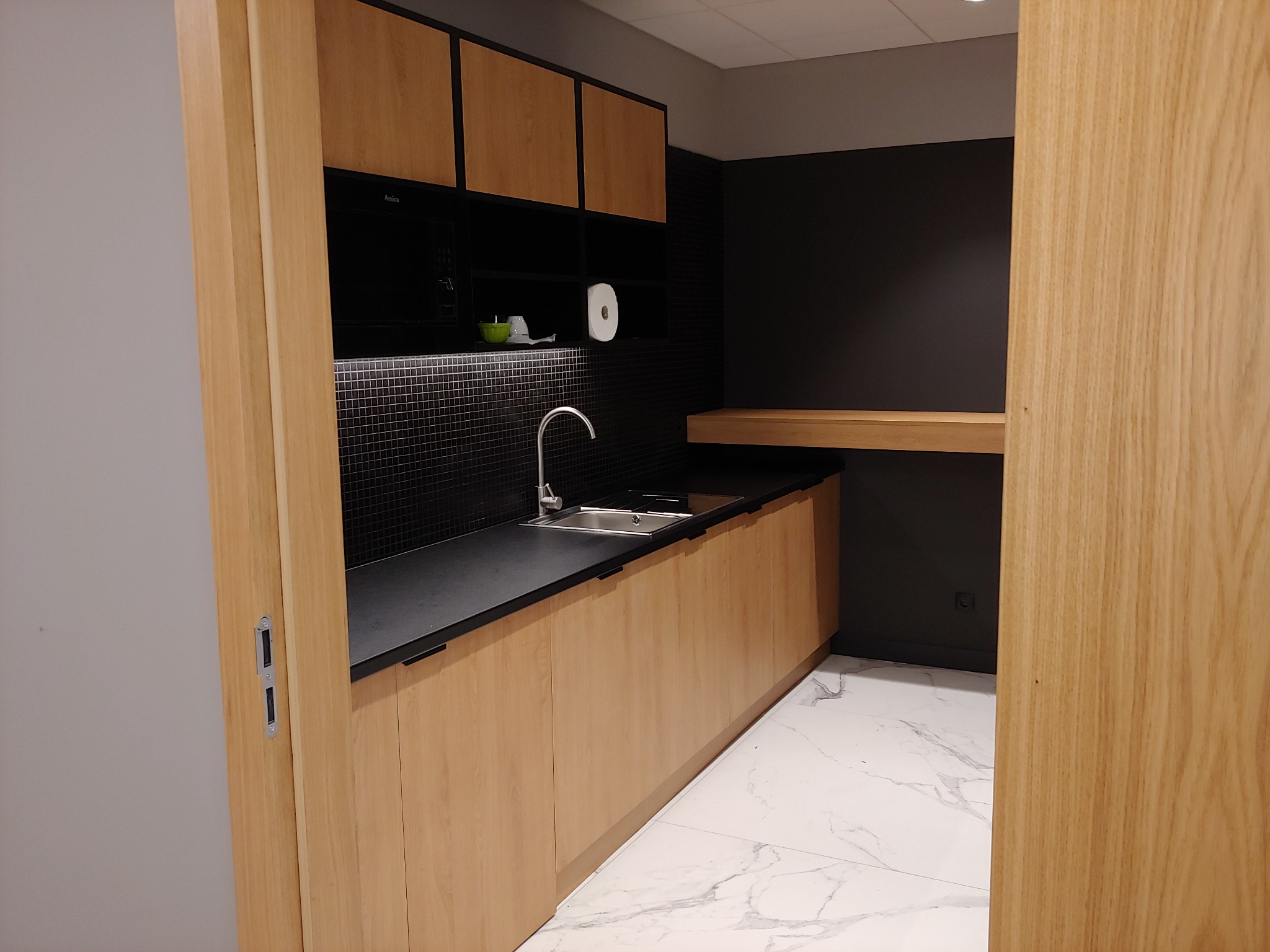 Nowoczesna kuchnia biurowa z drewnianymi szafkami, czarnym blatem i mozaiką, widok zza otwartych drzwi.