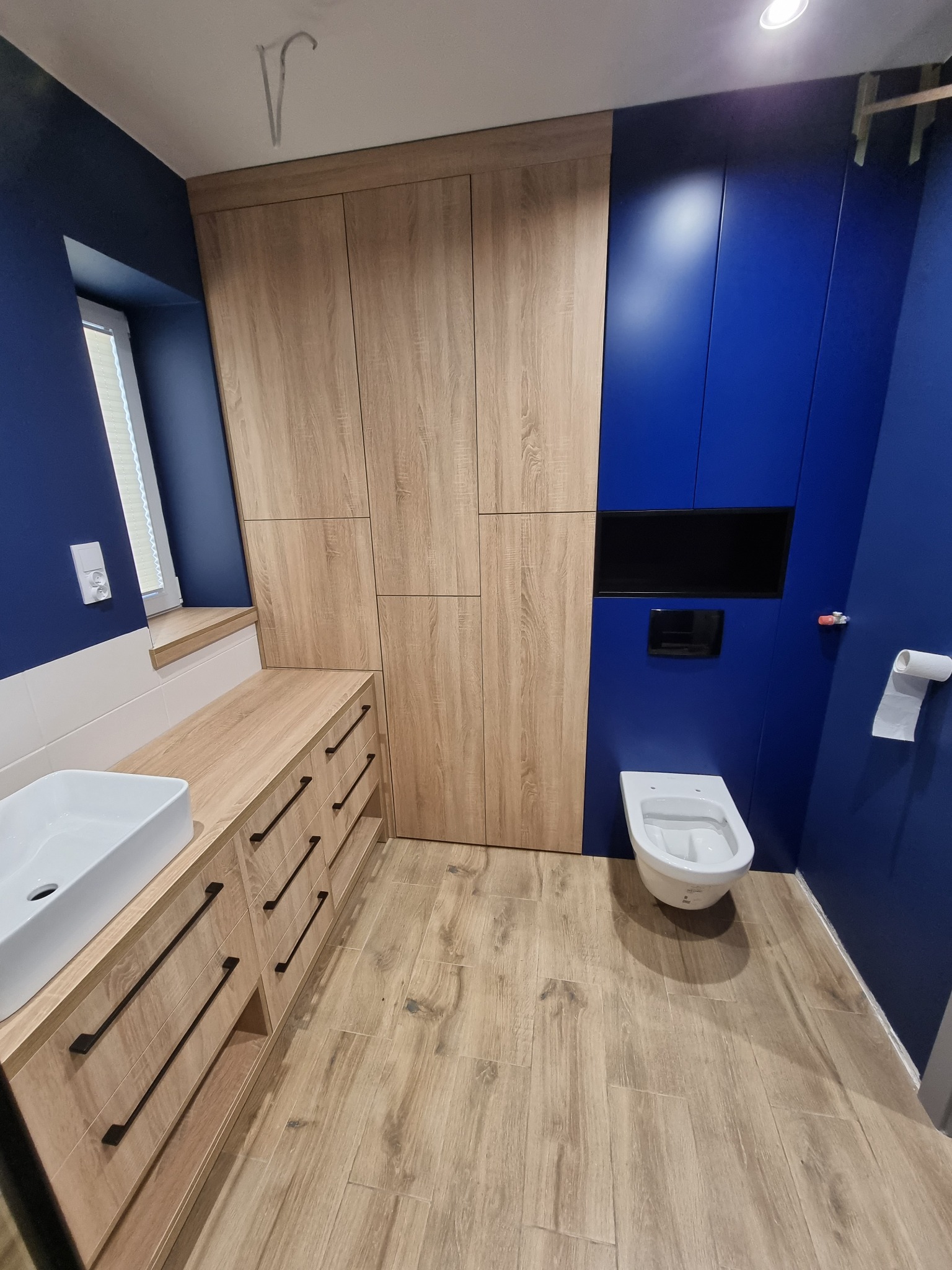 Nowoczesna łazienka z szafkami z drewnopodobnymi frontami, umywalką nablatową, toaletą podwieszaną i ścianami w kolorze głębokiego granatu.