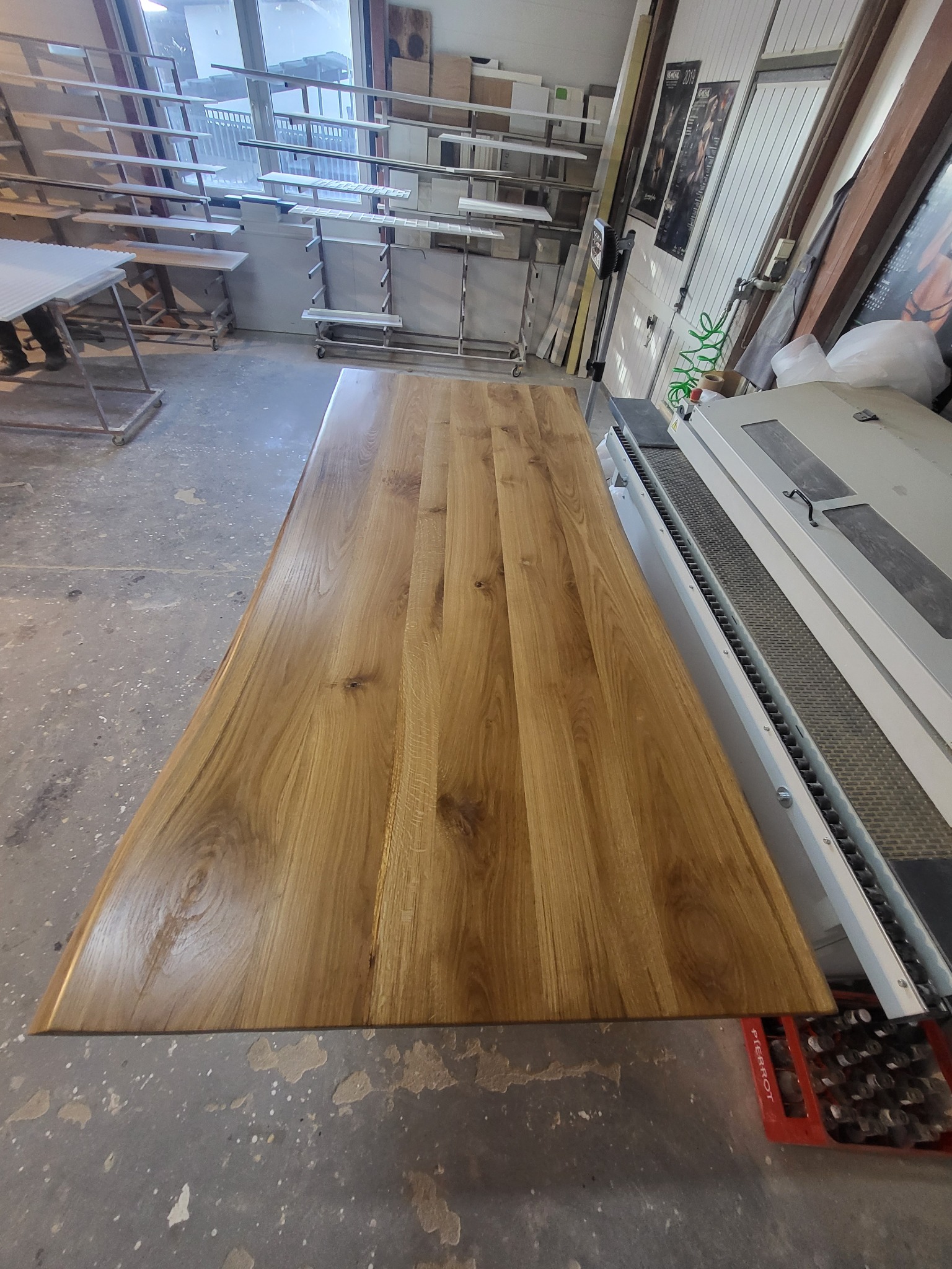 Polerowany blat z drewna dębowego na linii produkcyjnej, widoczne słoje i naturalna kolorystyka drewna, w tle regały z elementami do mebli.