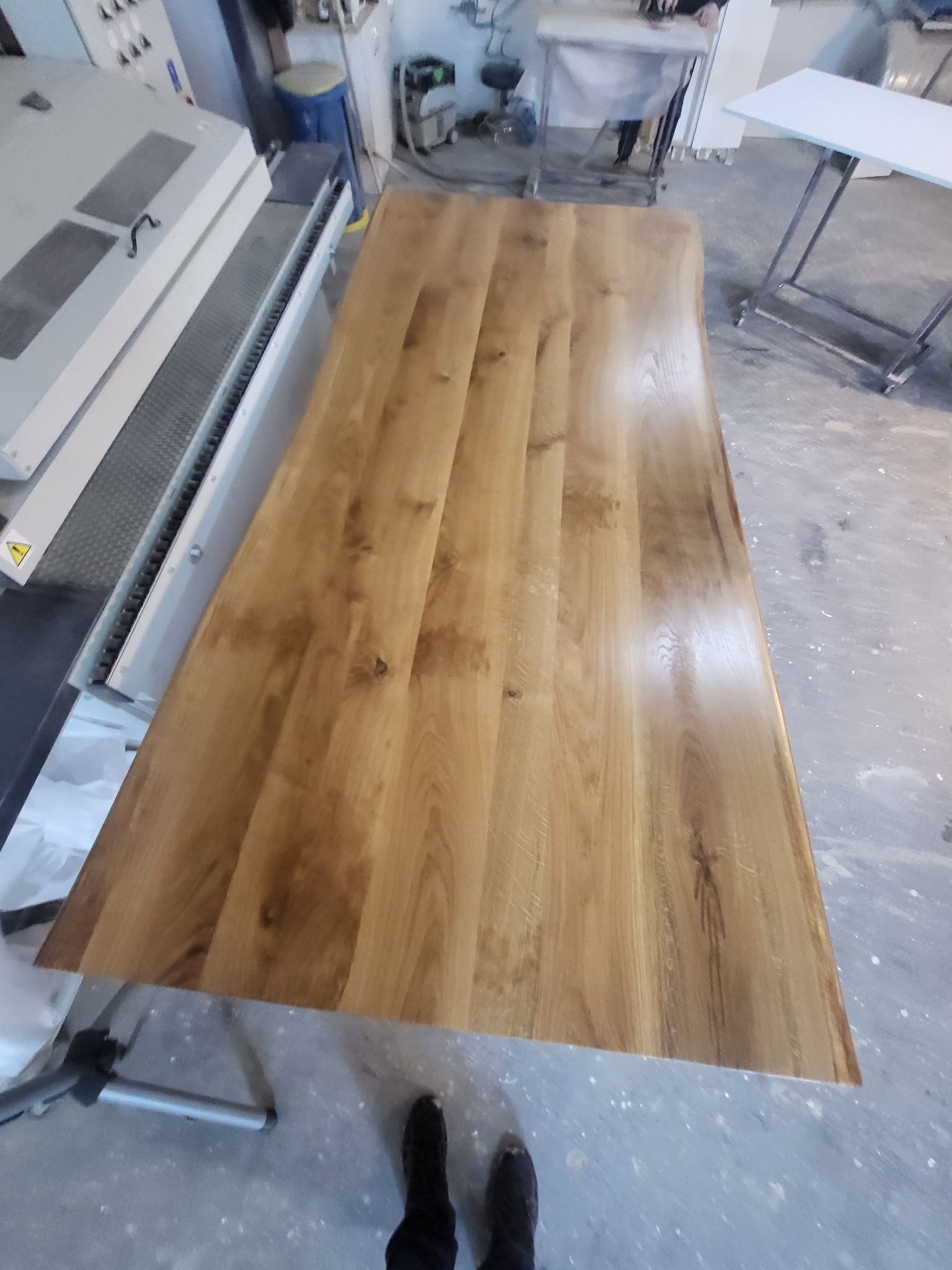 Blat z jasnego drewna leżący na stole warsztatowym, widoczne słoje drewna, obok maszyna do obróbki drewna, na podłodze ślady farby.