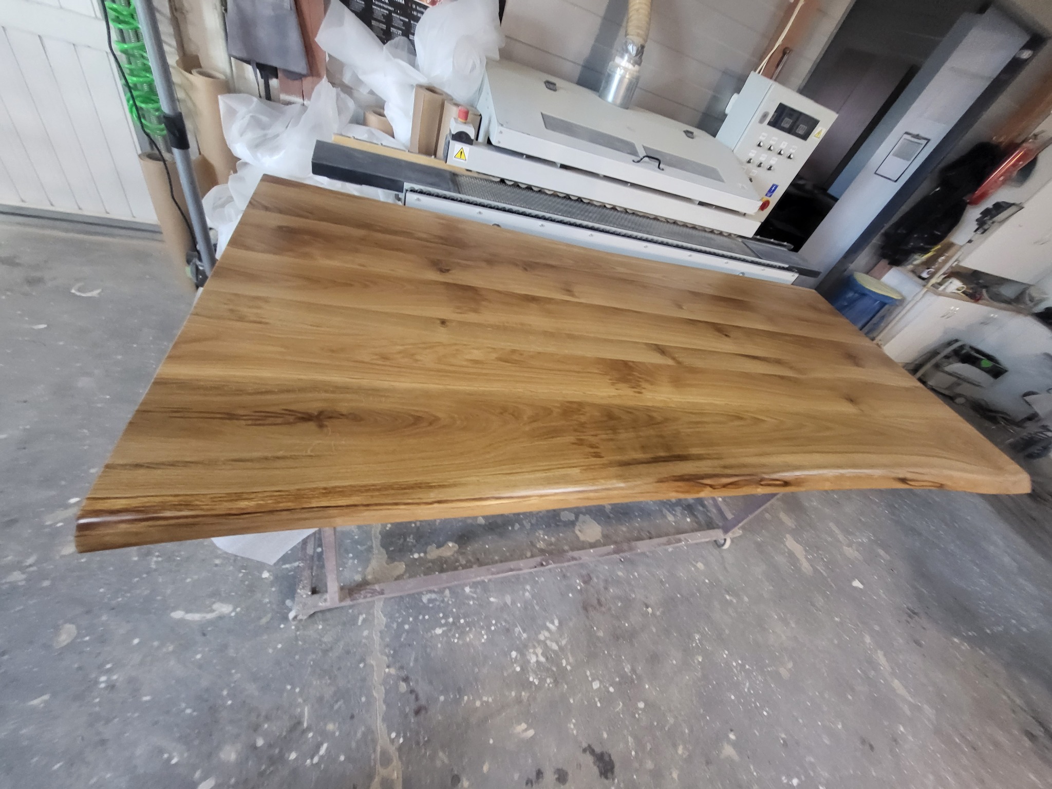 Obróbka blatu drewnianego na metalowym stelażu, widoczna maszyna szlifująca i materiały pomocnicze w tle warsztatu.