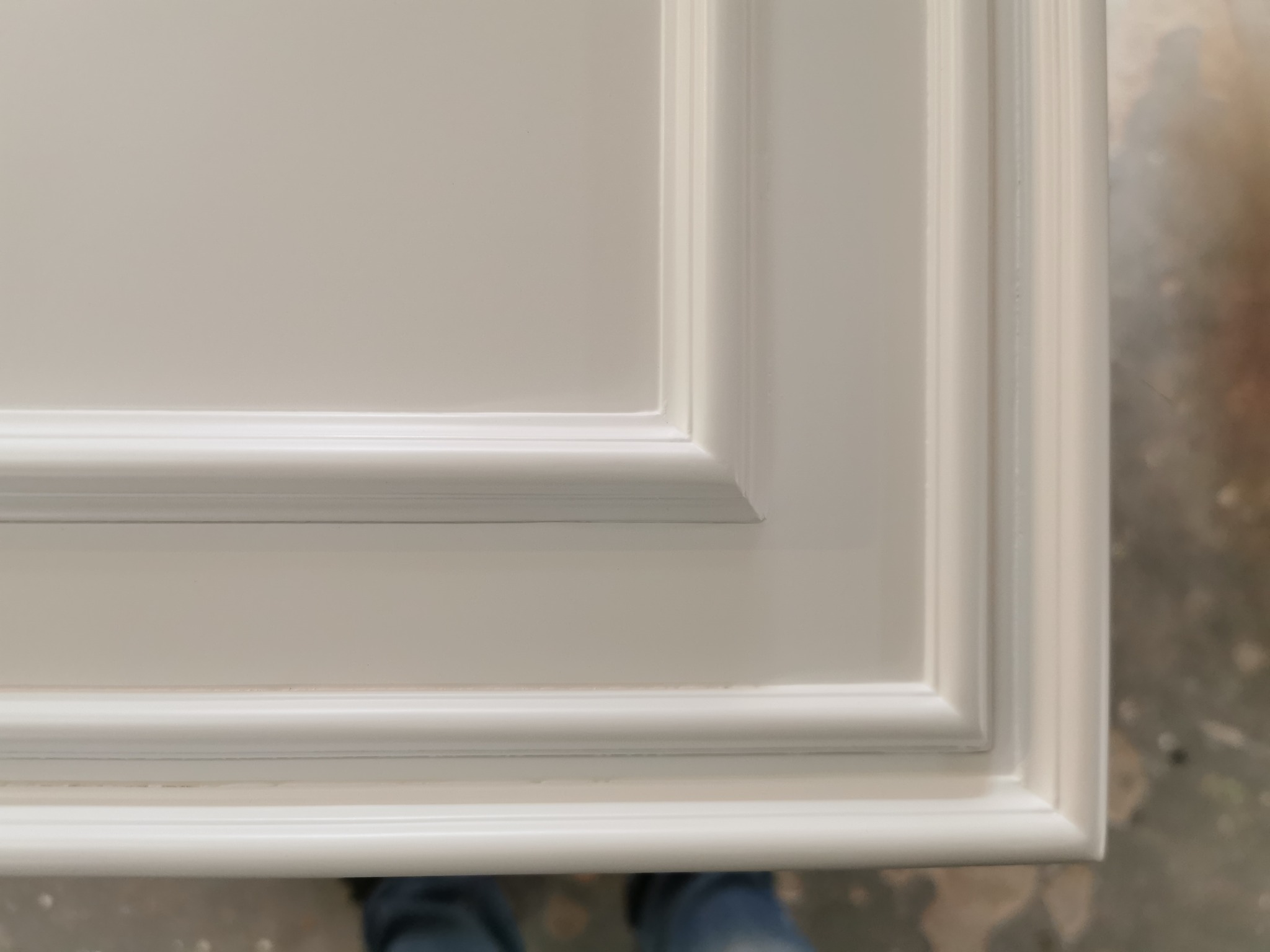 Fragment białego, frezowanego frontu meblowego z widocznymi detalami wykończenia i fragmentem podłogi w tle.