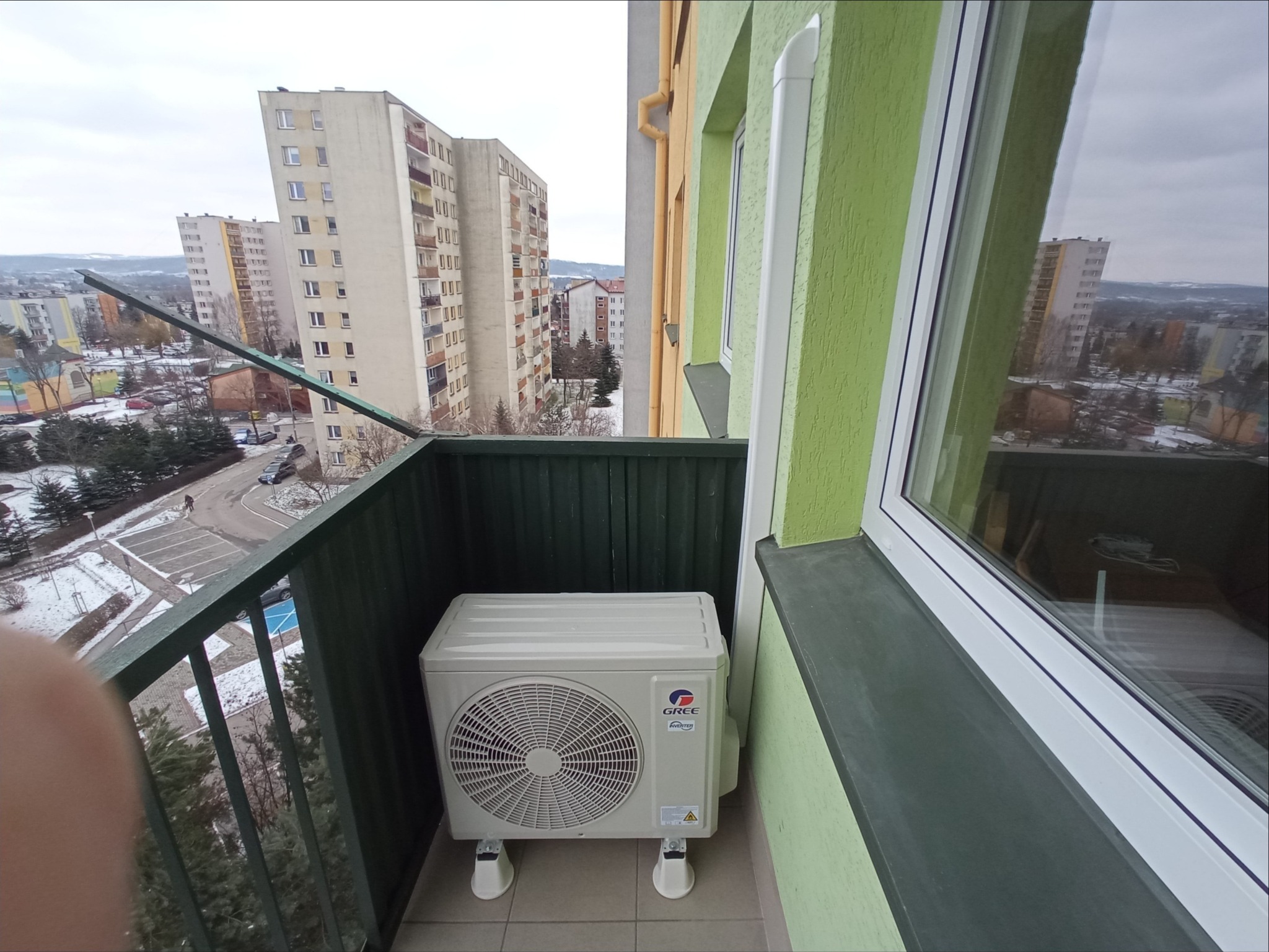 Zewnętrzna jednostka klimatyzacji marki Gree zamontowana na balkonie mieszkania w bloku, widok na osiedle miejskie w pochmurny dzień.