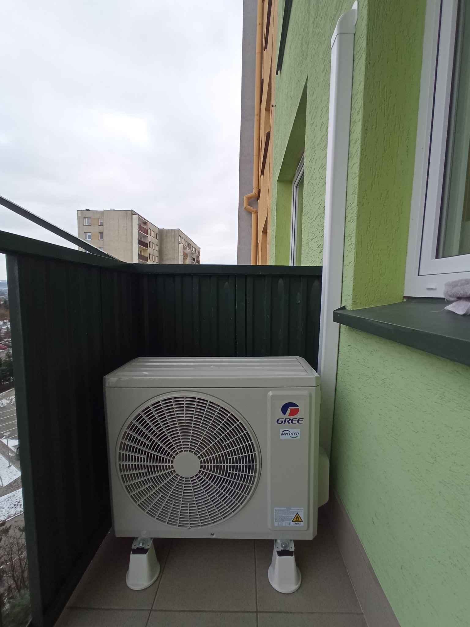 Zewnętrzna jednostka klimatyzacji marki Gree, zamontowana na balkonie bloku mieszkalnego z widokiem na szary krajobraz miejski w pochmurny dzień.