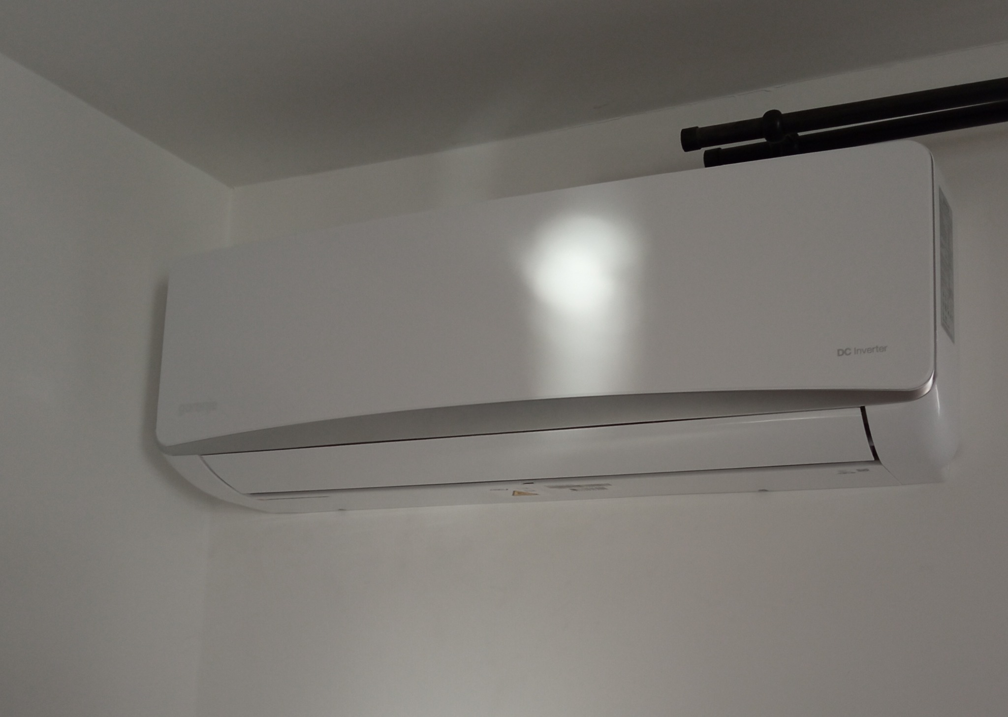 Biała jednostka wewnętrzna klimatyzacji Gorenje DC Inverter zamontowana na ścianie w jasnym pomieszczeniu, widoczne czarne rury instalacyjne pod sufitem.