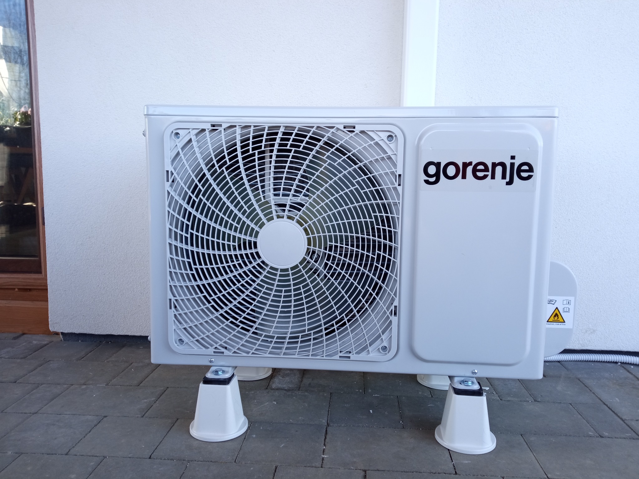 Zewnętrzna jednostka klimatyzacji Gorenje na szarym bruku przy białej ścianie budynku, widoczny znak ostrzegawczy o ryzyku pożaru.