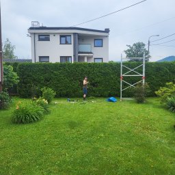 Hermes GardenBud - Mężczyzna przy wysokim żywopłocie, obok rozłożone narzędzia ogrodnicze i rusztowanie. W tle dom z balkonem i zielony trawnik z kwiatami.