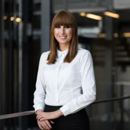 Portret uśmiechniętej adwokatki Alicji Krzyśko-Makulskiej w białej koszuli i czarnej spódnicy, opierającej się o metalową barierkę na tle nowoczesnego biura.