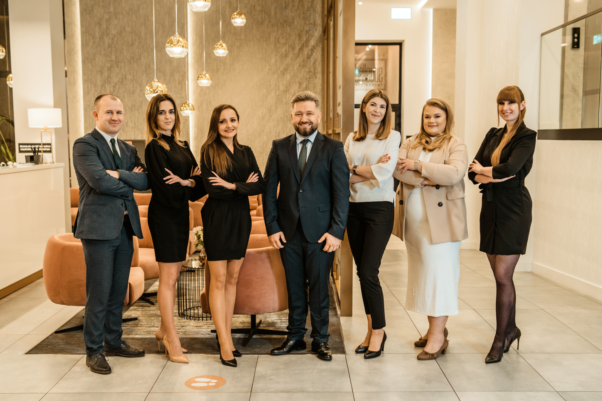 Grupa sześciu prawników, trzech mężczyzn i trzy kobiety, stojących ramię w ramię w nowoczesnym wnętrzu biura z dekoracyjnym oświetleniem i fotelami w tle. Wszyscy ubrani w formalne stroje biznesowe...