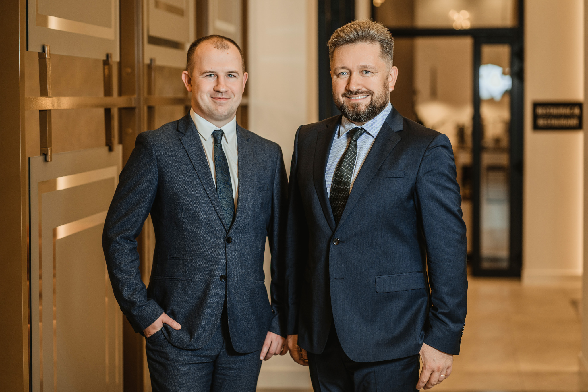 Portret dwóch uśmiechniętych adwokatów w garniturach, stojących obok siebie w eleganckim wnętrzu kancelarii z dekoracyjnymi panelami ściennymi i szklanymi drzwiami w tle.