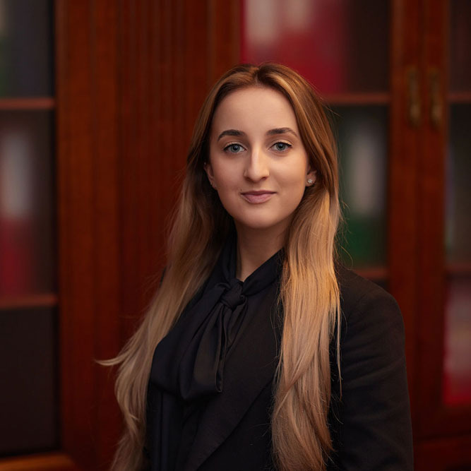 Portret młodej kobiety, asystent adwokata, w czarnym żakiecie i bluzce z wiązaniem pod szyją, na tle drewnianej biblioteczki z czerwonymi akcentami.